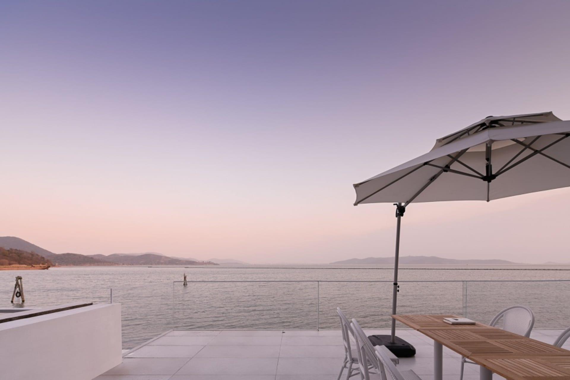 Modern dining area featuring a glass table and chairs, overlooking a serene lake at sunset.