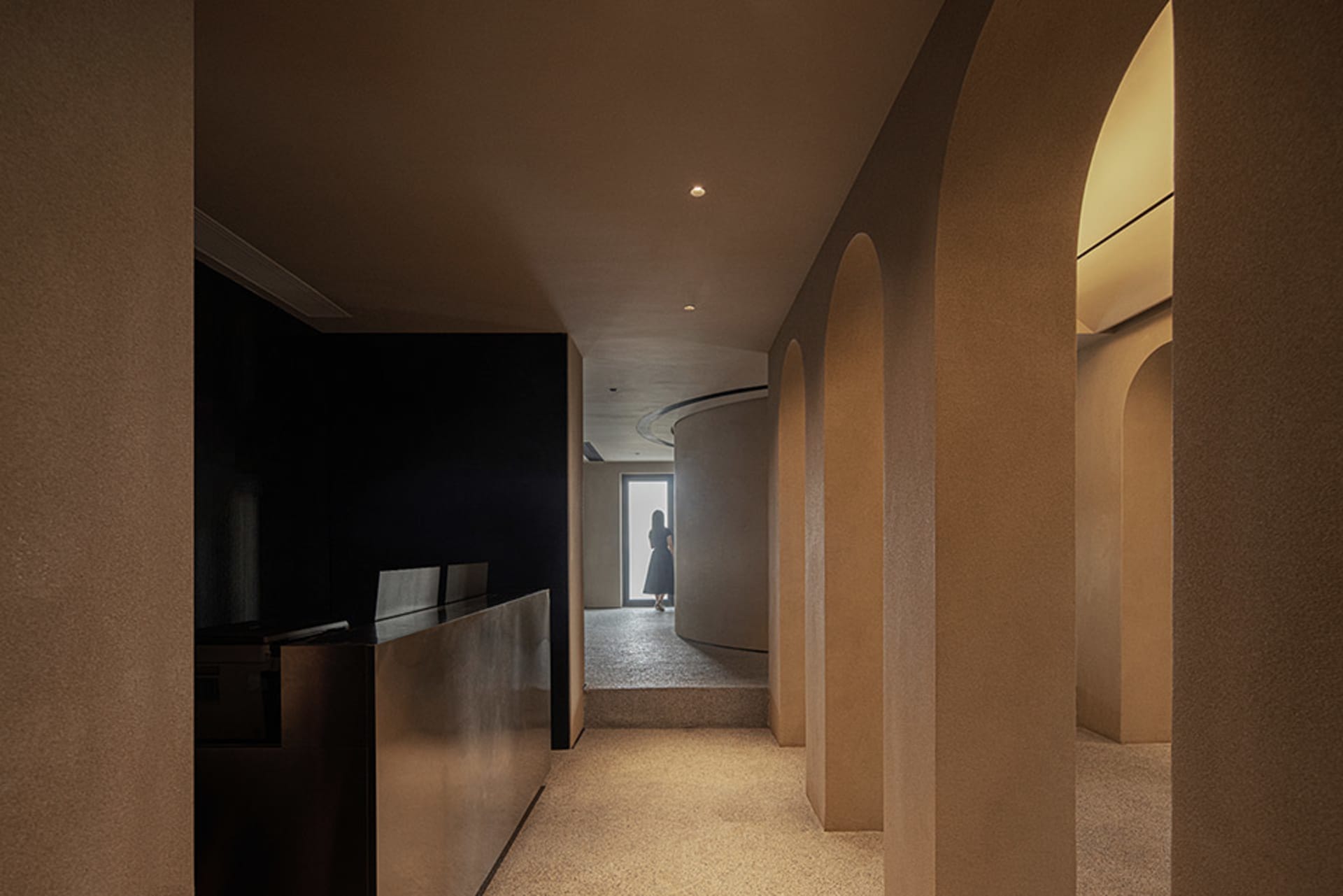 A modern architectural corridor with arched openings and concrete walls, featuring a person standing in the distance.