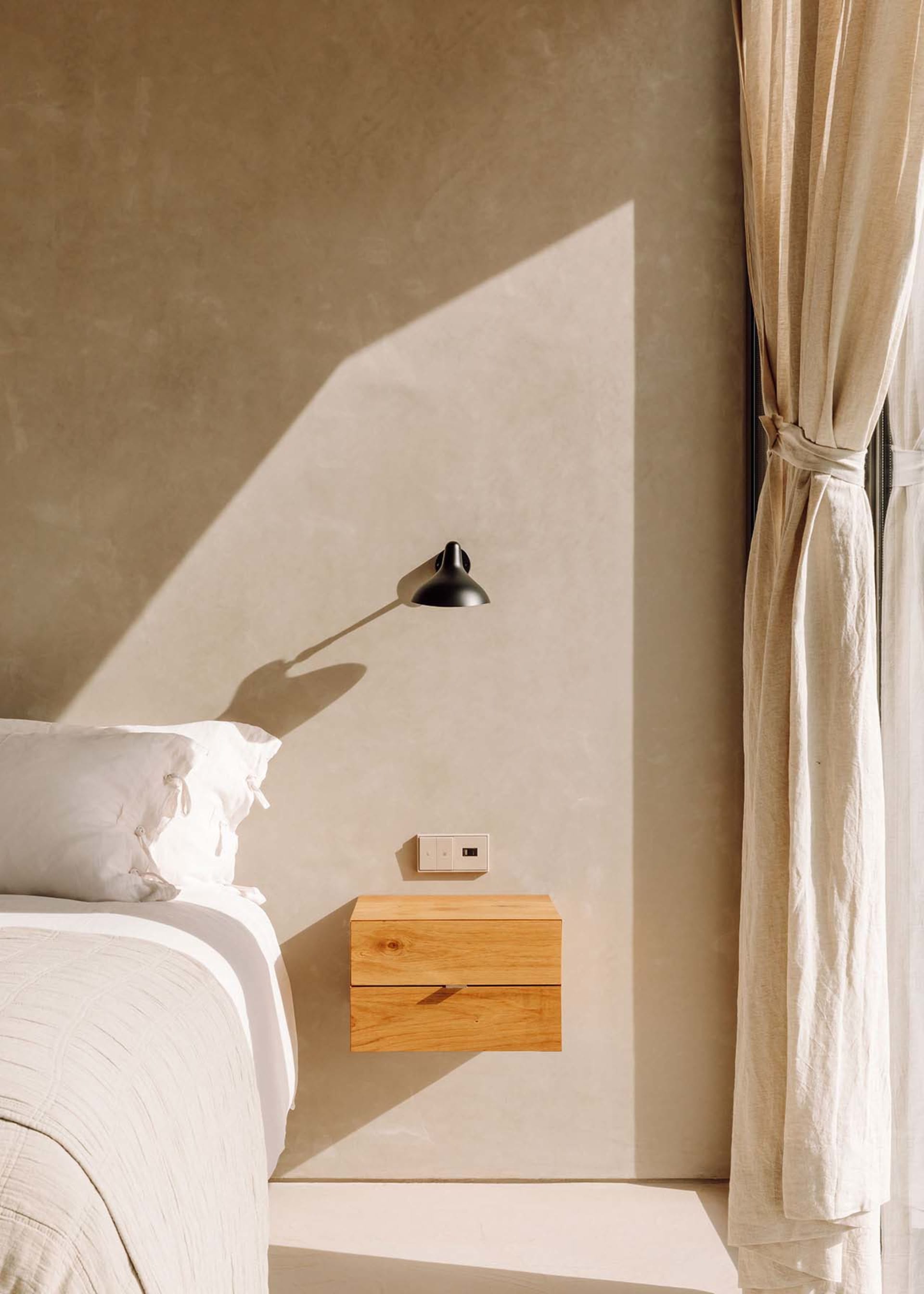 Minimalist bedroom with a wooden floating nightstand, window with curtains, and soft lighting from a wall-mounted sconce.