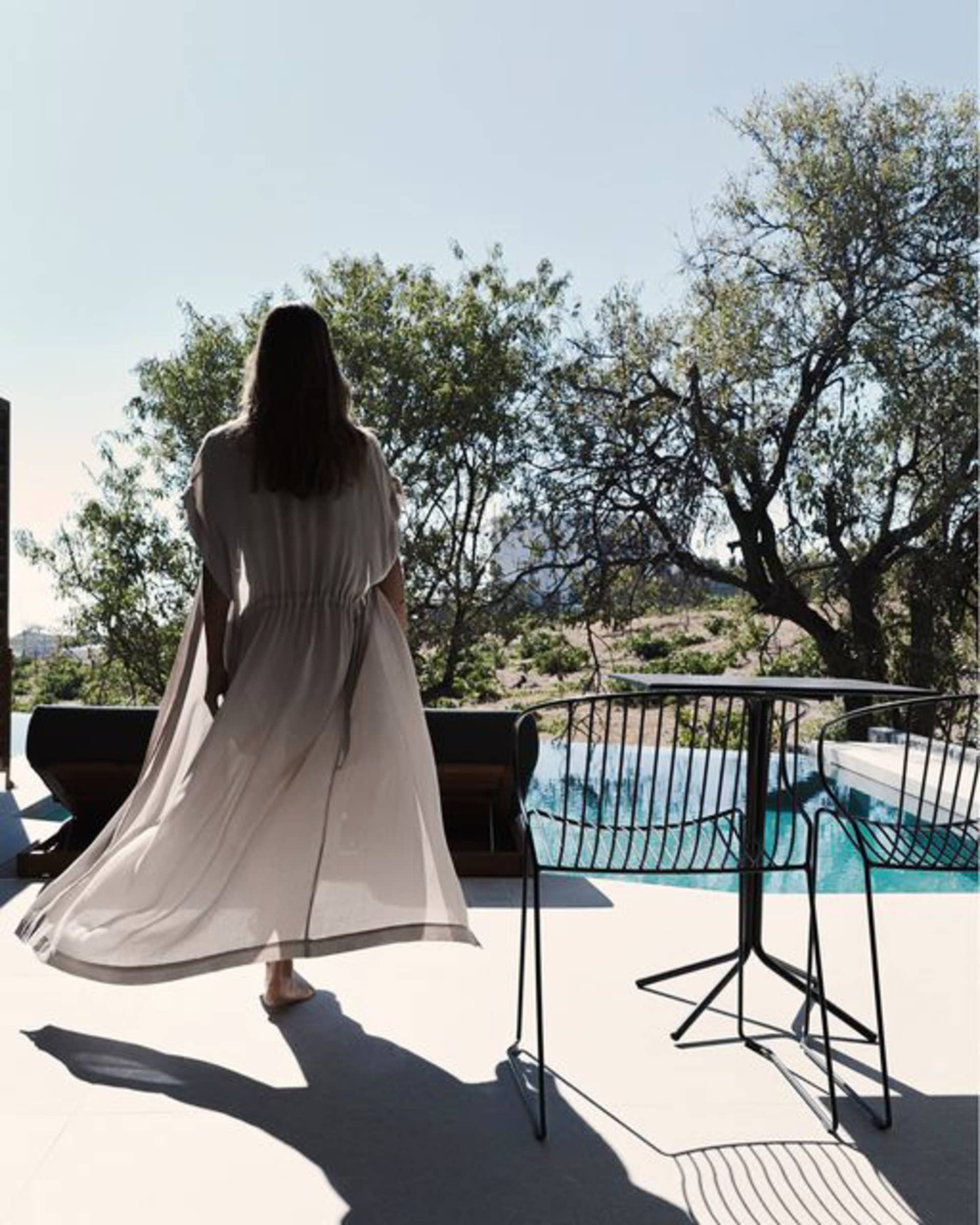 A woman in a long dress walks on a patio with concrete, metal, and glass elements, overlooking a pool and trees.