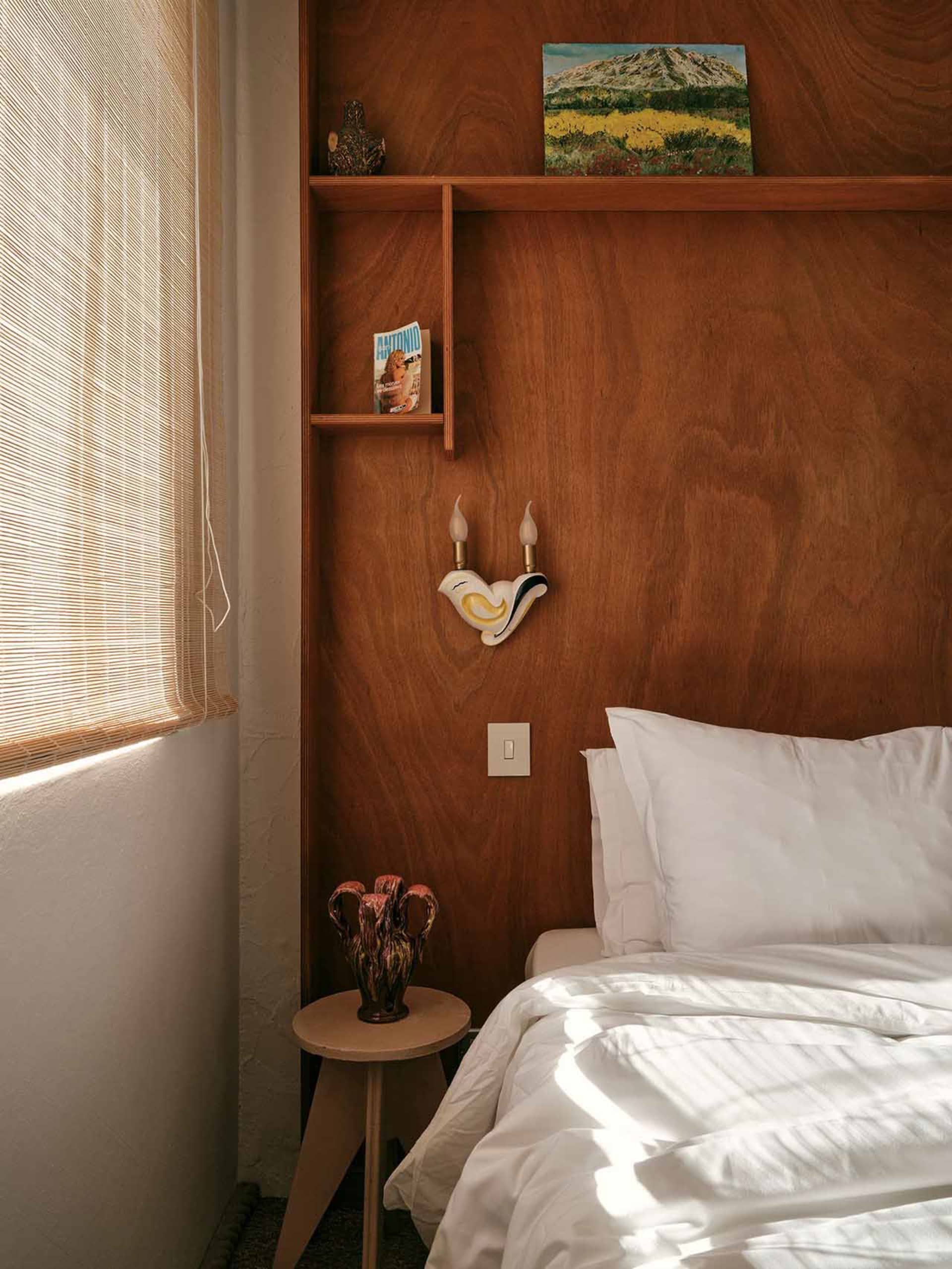 A minimalist bedroom features a bed with white bedding, a wooden headboard, and a wall-mounted sconce. The room is adorned wi