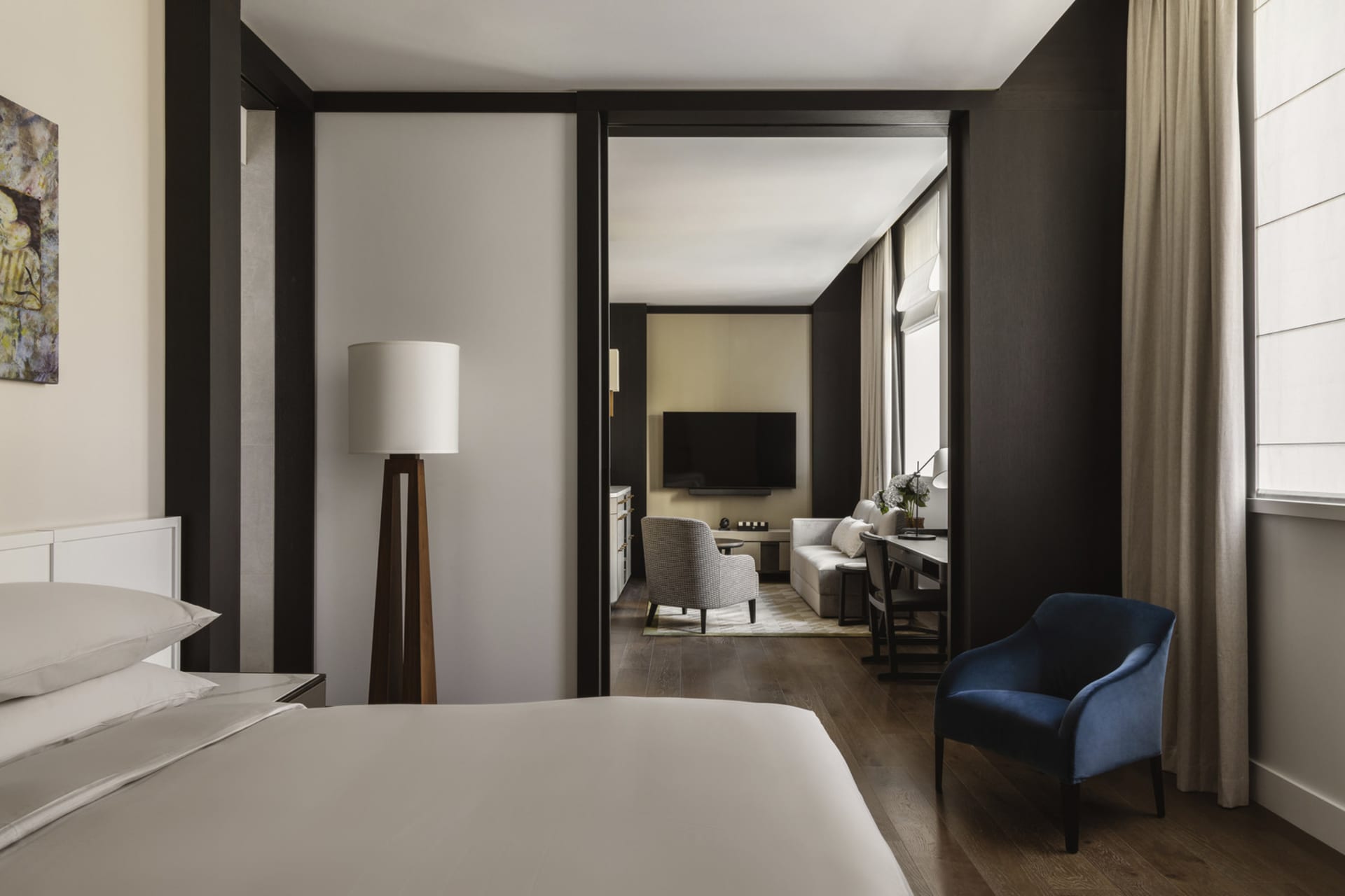 A modern hotel room featuring a minimalist bedroom with a bed, lamp, and a door leading to a living space.