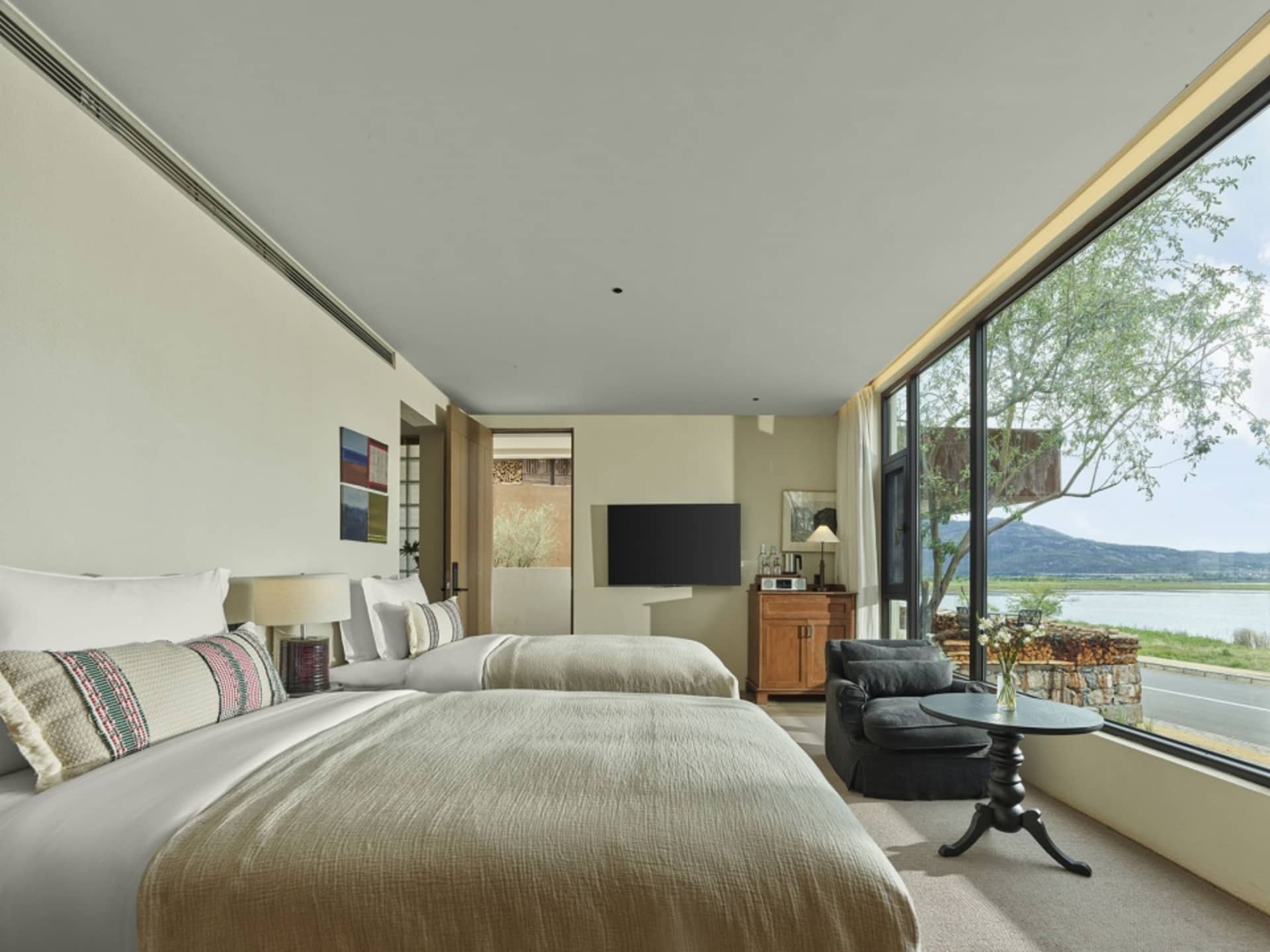 A modern minimalist bedroom with two twin beds, a flat-screen TV, and a view of the water.