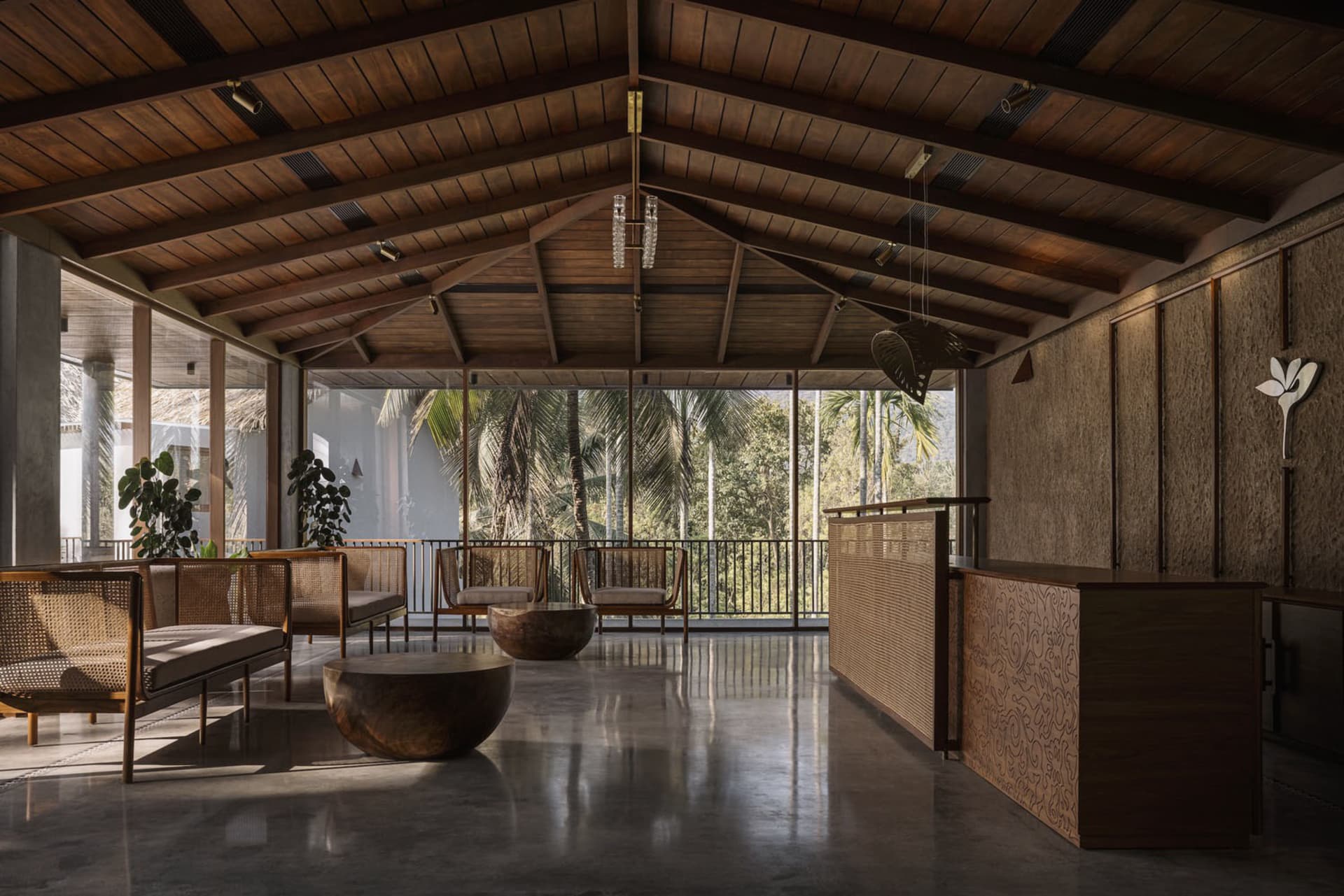 A modern minimalist lobby with wooden furniture, concrete flooring, and a view of palm trees through large windows.