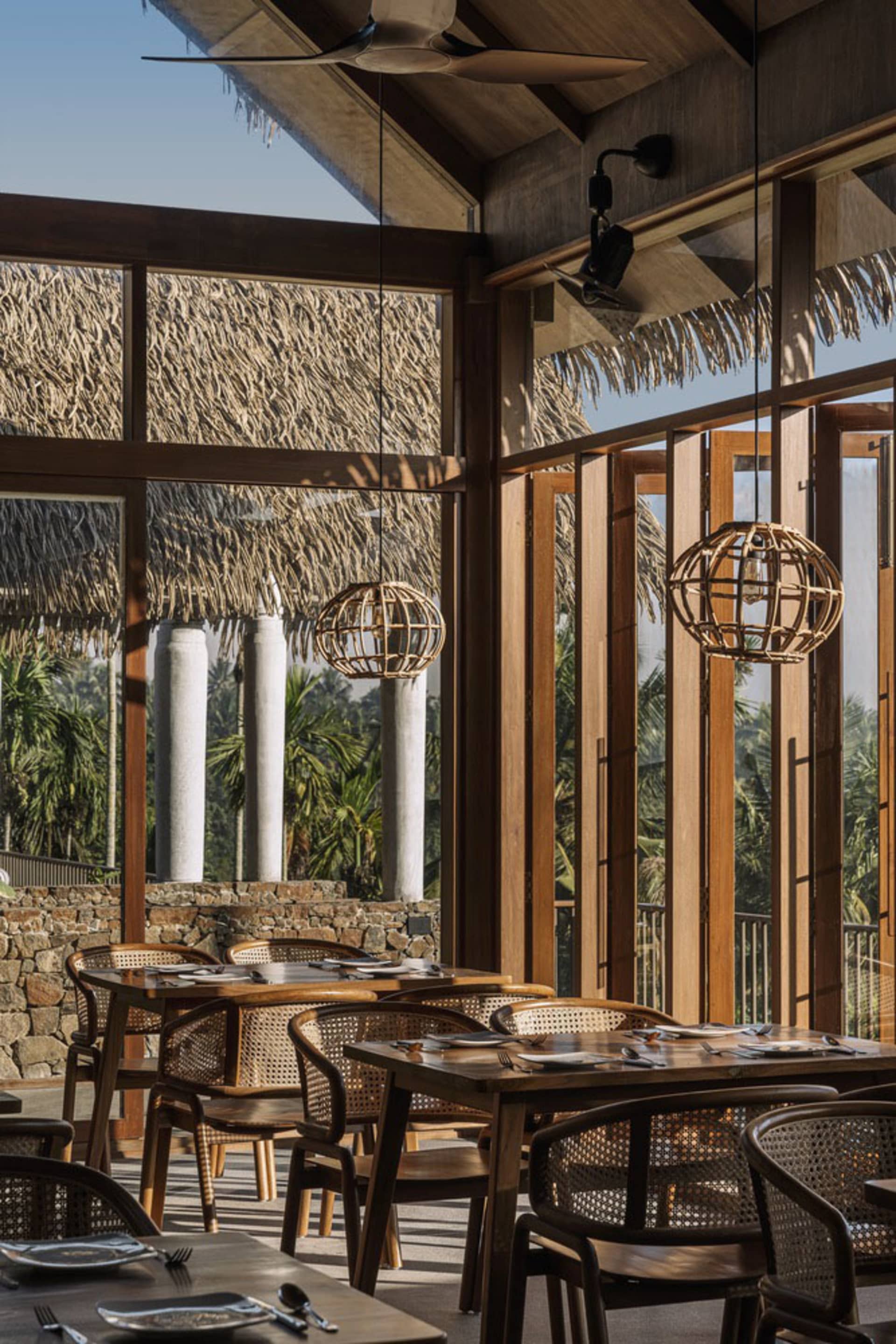 A restaurant interior with natural wood furniture and large windows showcasing a tropical outdoor setting.