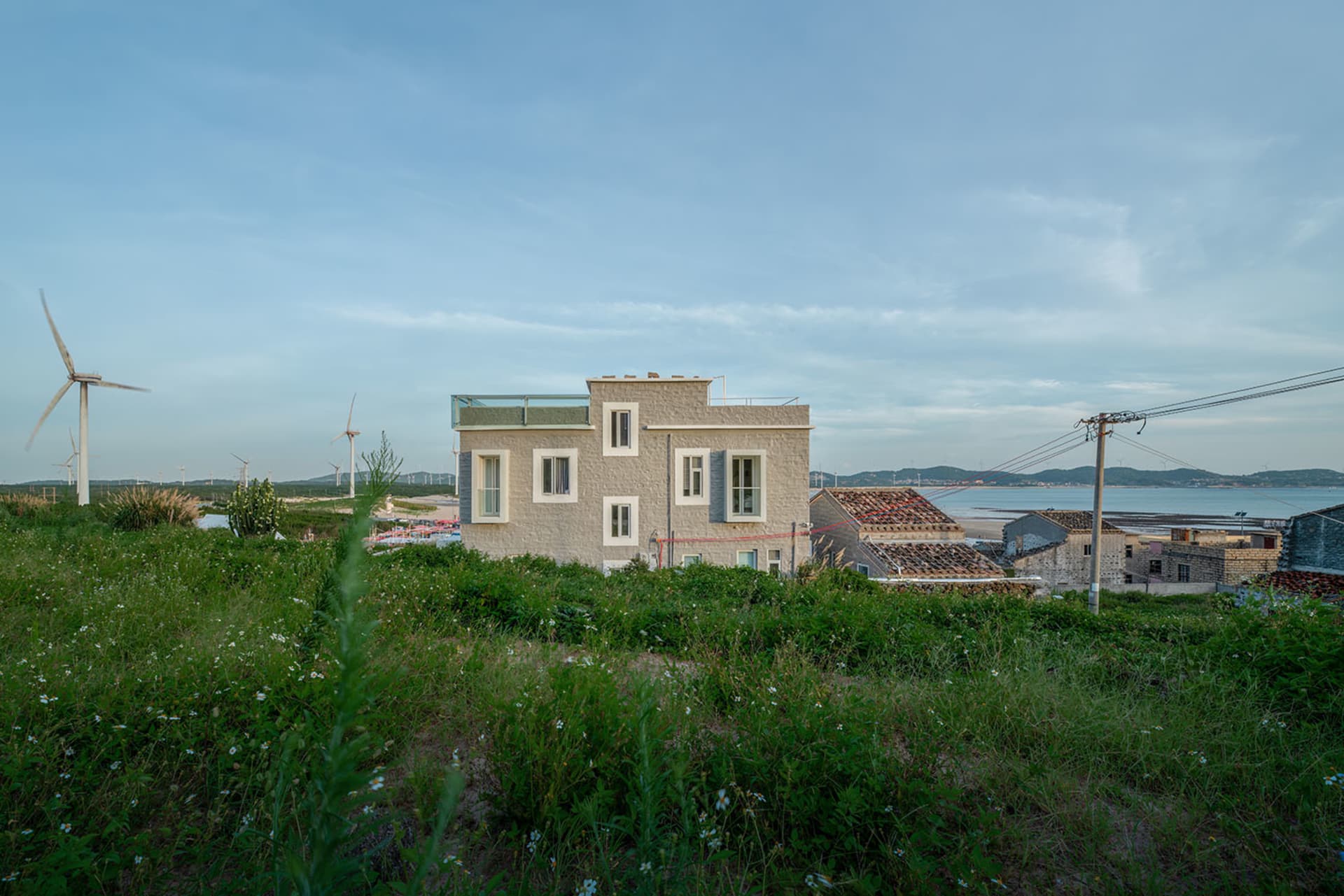A minimalist-style beach house with concrete walls, surrounded by greenery and wind turbines.