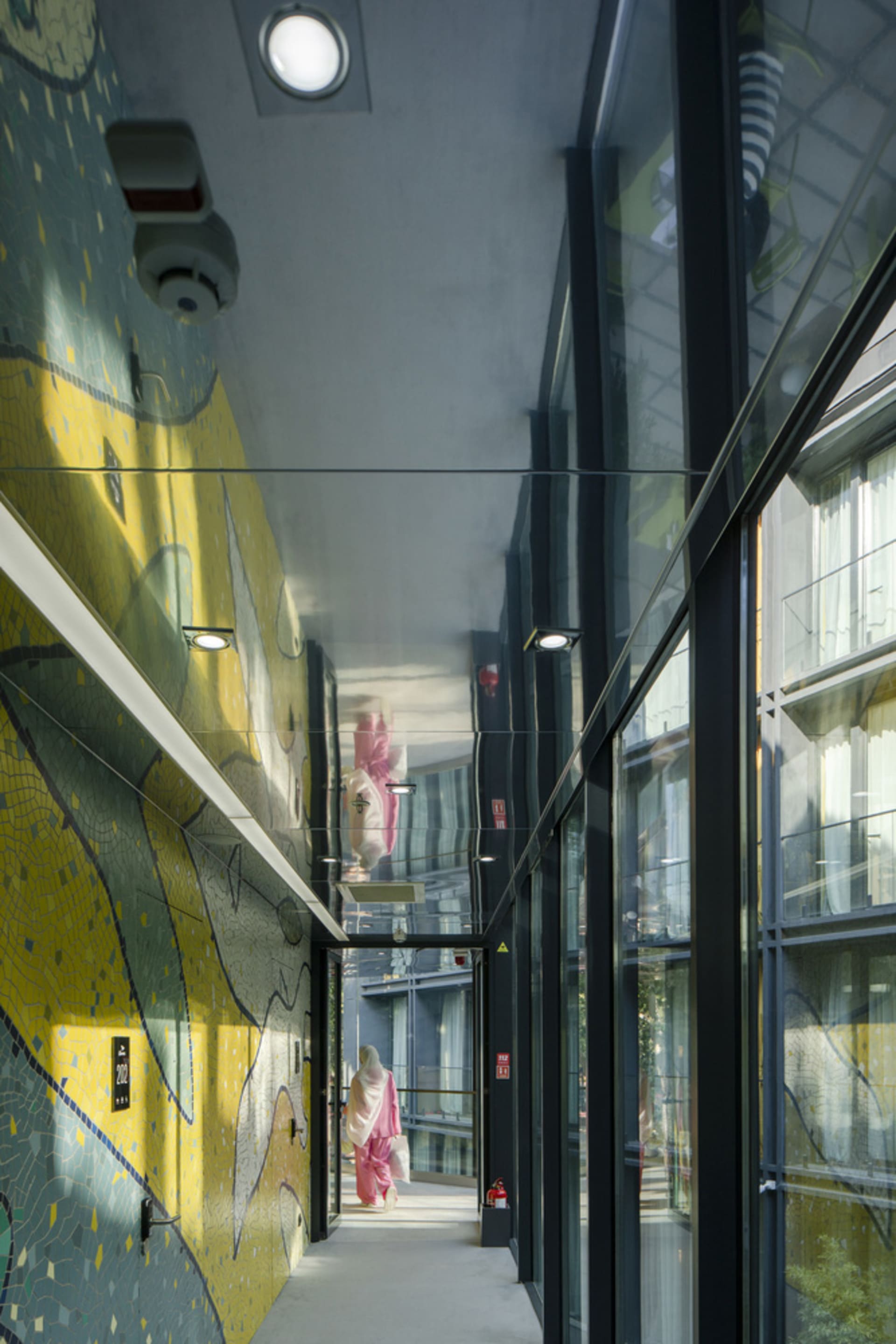 A modern hallway with glass and metal walls, featuring colorful wall art and a person in a pink outfit.