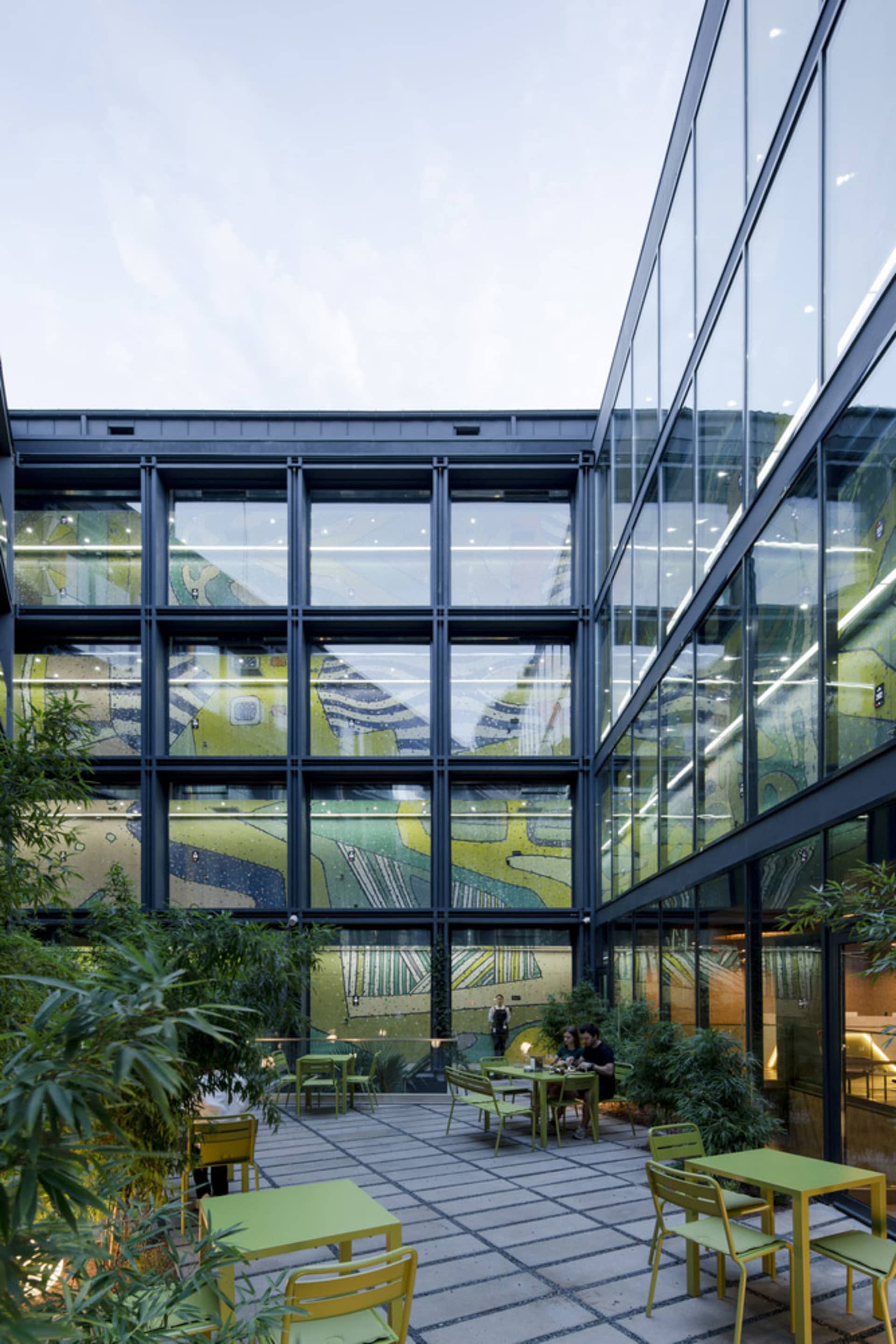 Outdoor patio area inside an office building with glass walls, featuring tables, chairs, and greenery.
