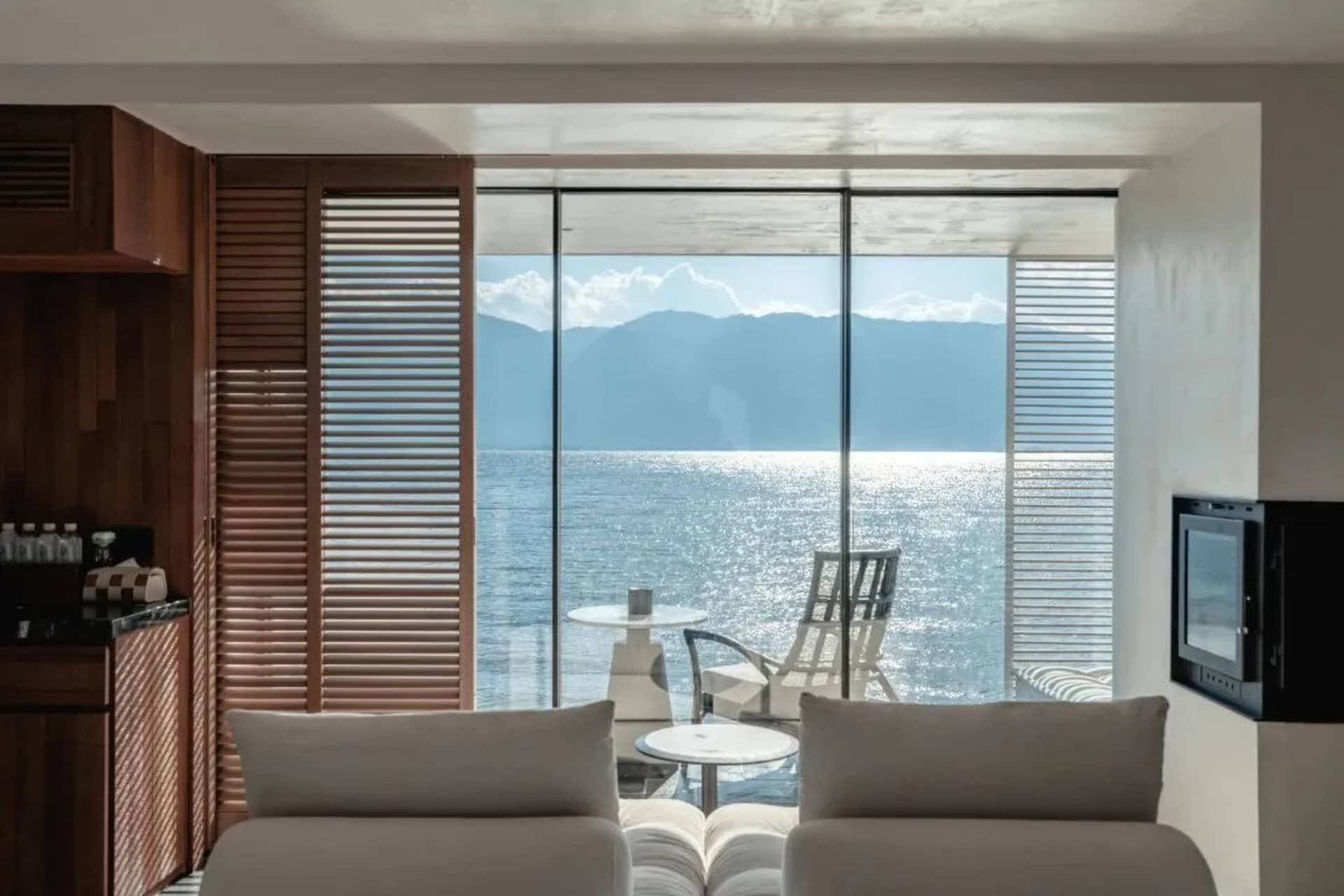 Contemporary living room overlooking the sea with wooden shutters, glass walls, and concrete furniture.
