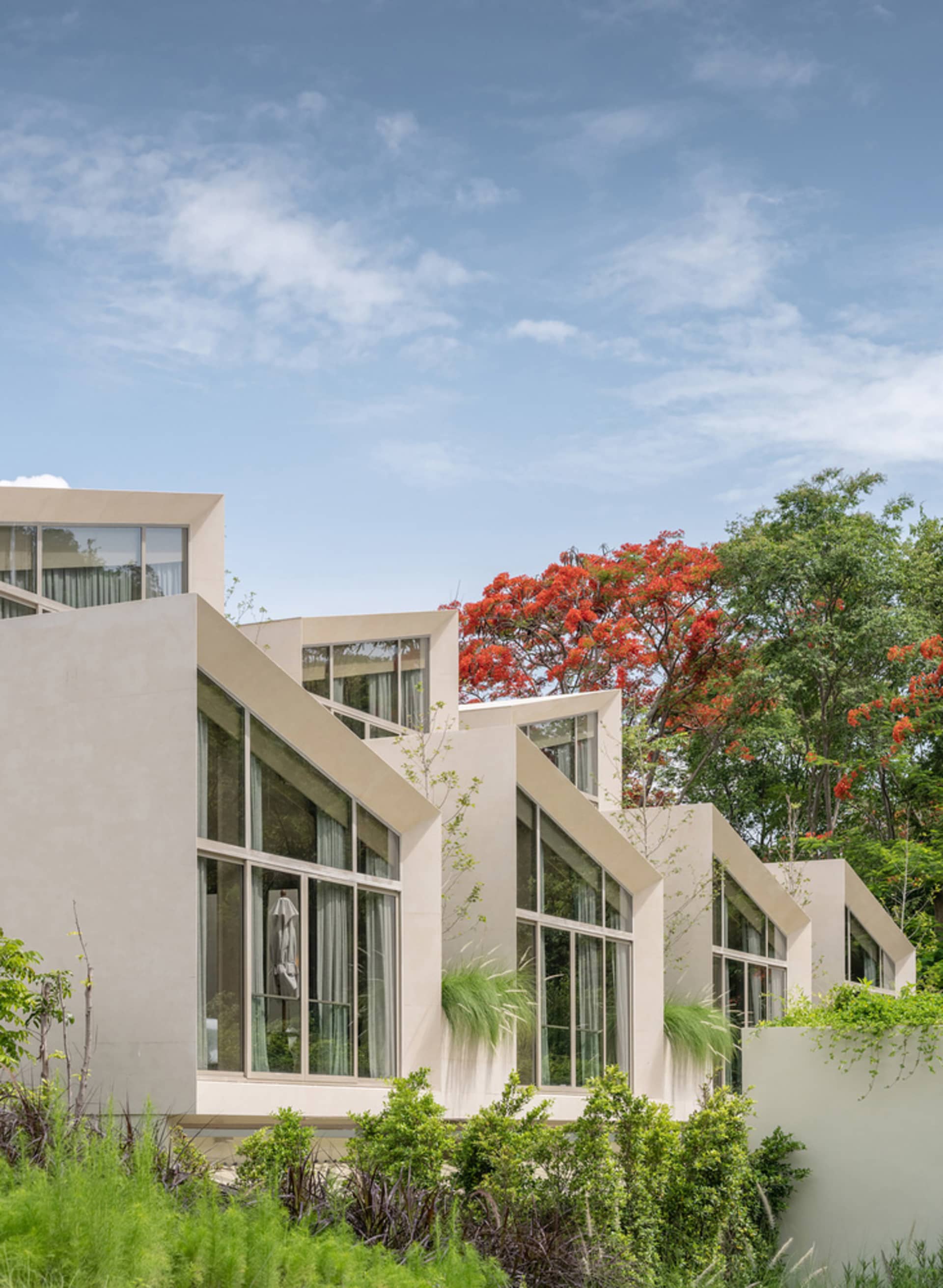 A modern house with large glass-paneled walls, a concrete foundation, and lush green plants.