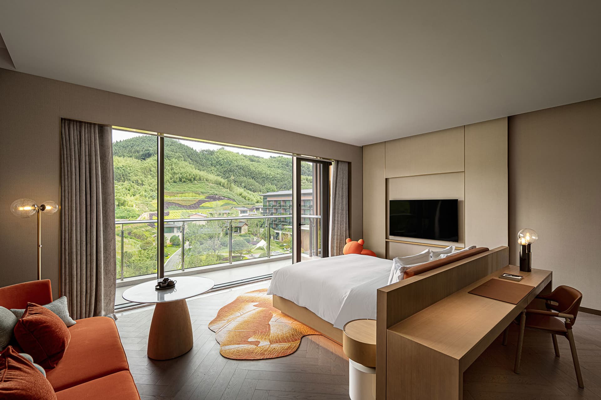 A modern residential bedroom featuring a glass window, a large bed, a wooden desk, and an orange lounge chair.