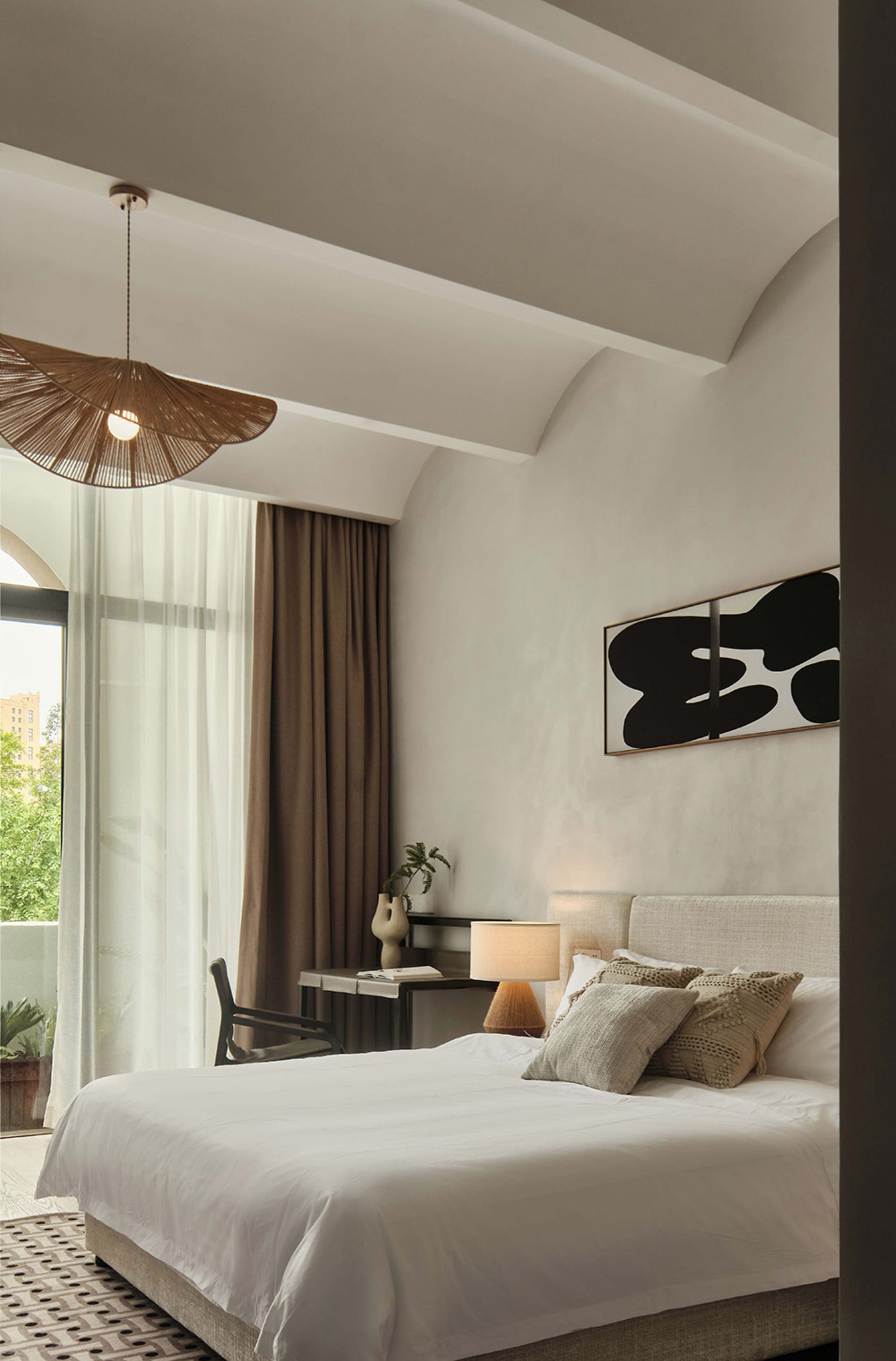 A minimalist bedroom featuring a bed with white linens, neutral-toned pillows, and a wicker pendant light hanging above it.