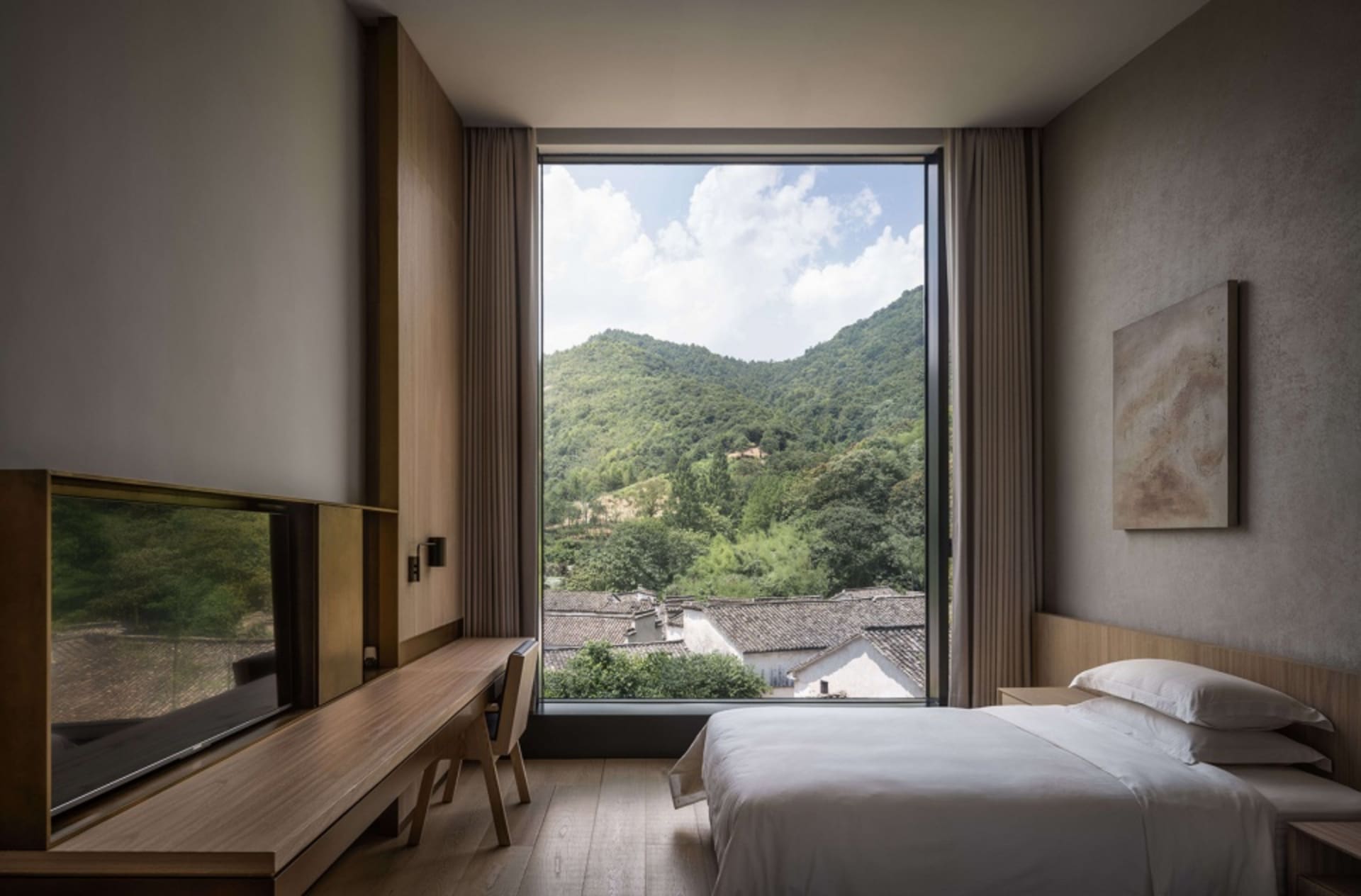 簡約臥室搭配白色家具與柔軟質感，設有大型窗戶可俯瞰綠意山景與屋頂