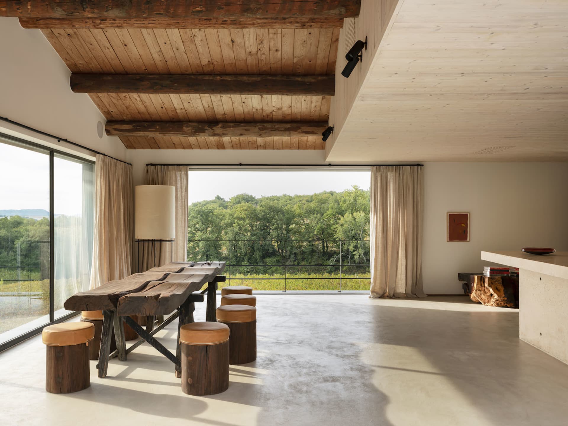 A modern minimalist living room with wooden accents and furniture, featuring large windows overlooking a forested area.