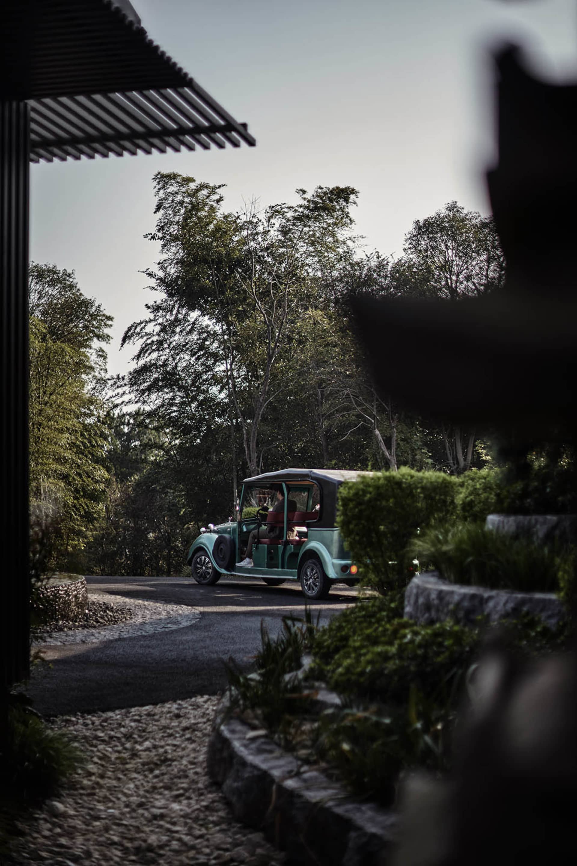 A blue vintage car driving on a cement road surrounded by lush greenery and trees.