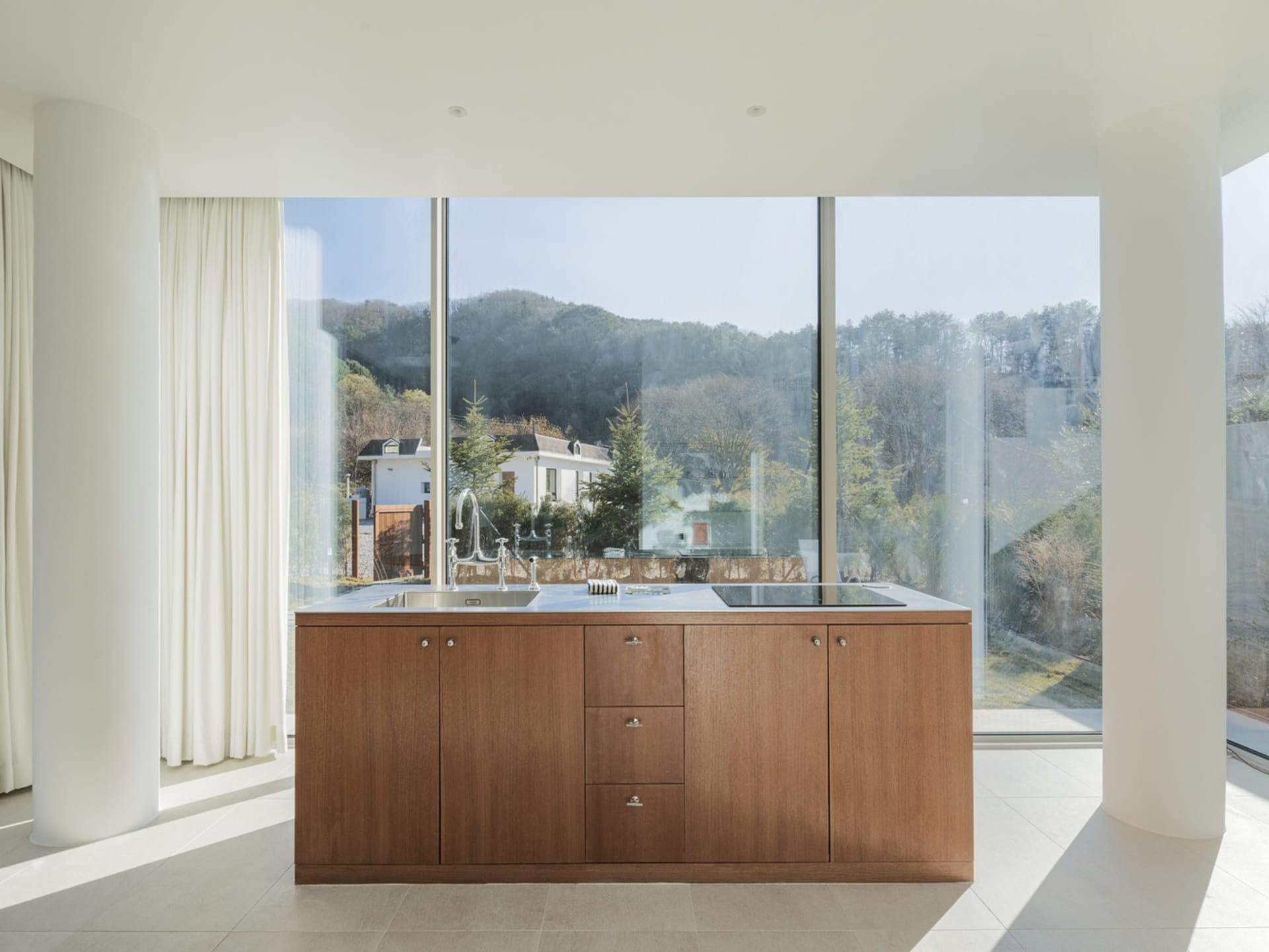 Modern, minimalist kitchen featuring wooden cabinetry and a large window overlooking trees and hills.