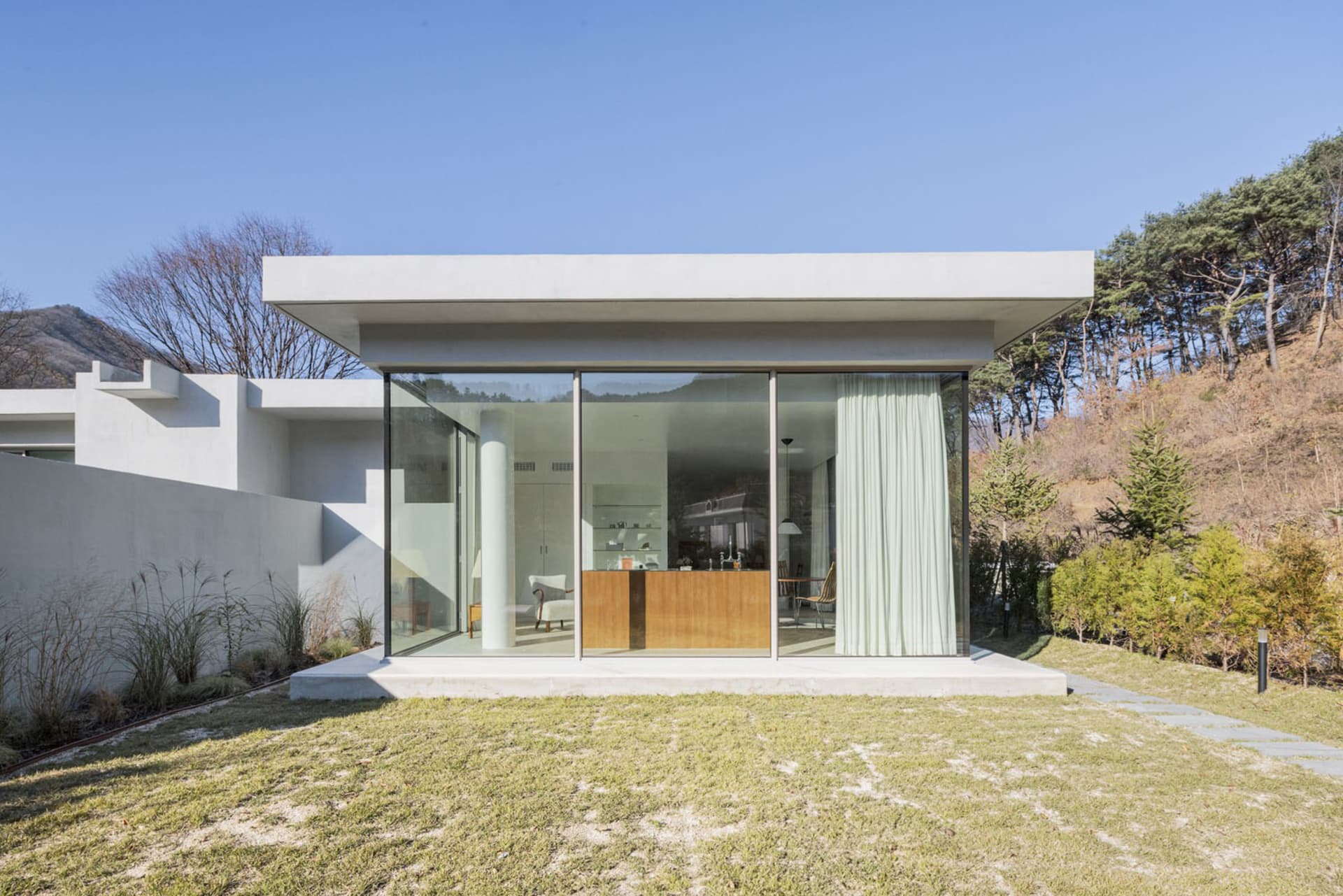 A minimalist modern house with large glass walls and a concrete base on a green hillside.