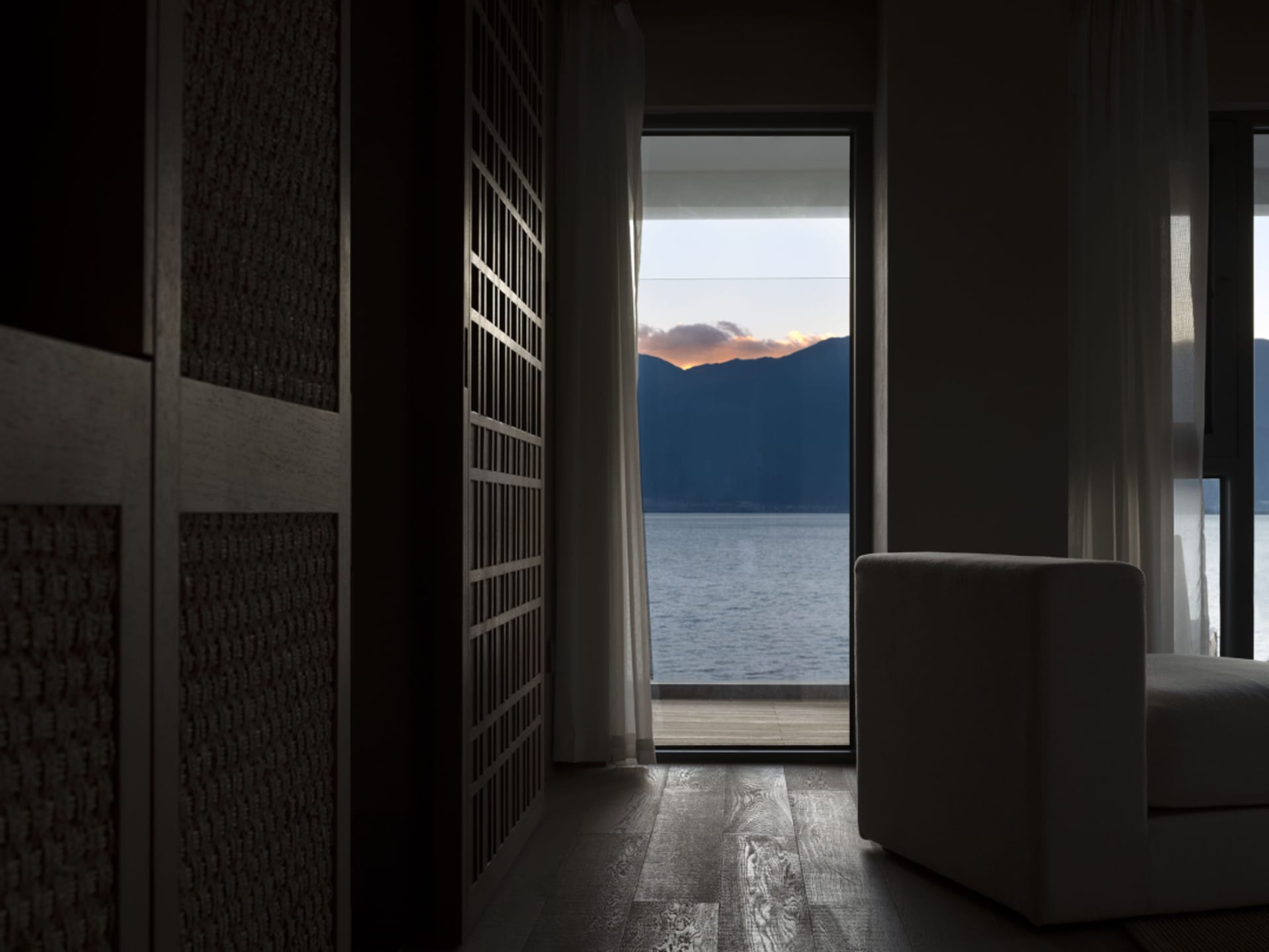 A cozy interior space with wooden floors and glass doors overlooking a stunning sunset over mountains and a body of water.