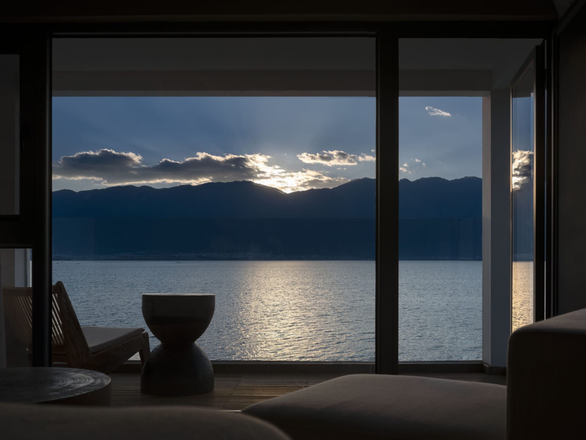 A serene view from a window overlooking the ocean at sunset, with mountains in the background and a wooden chair inside the r