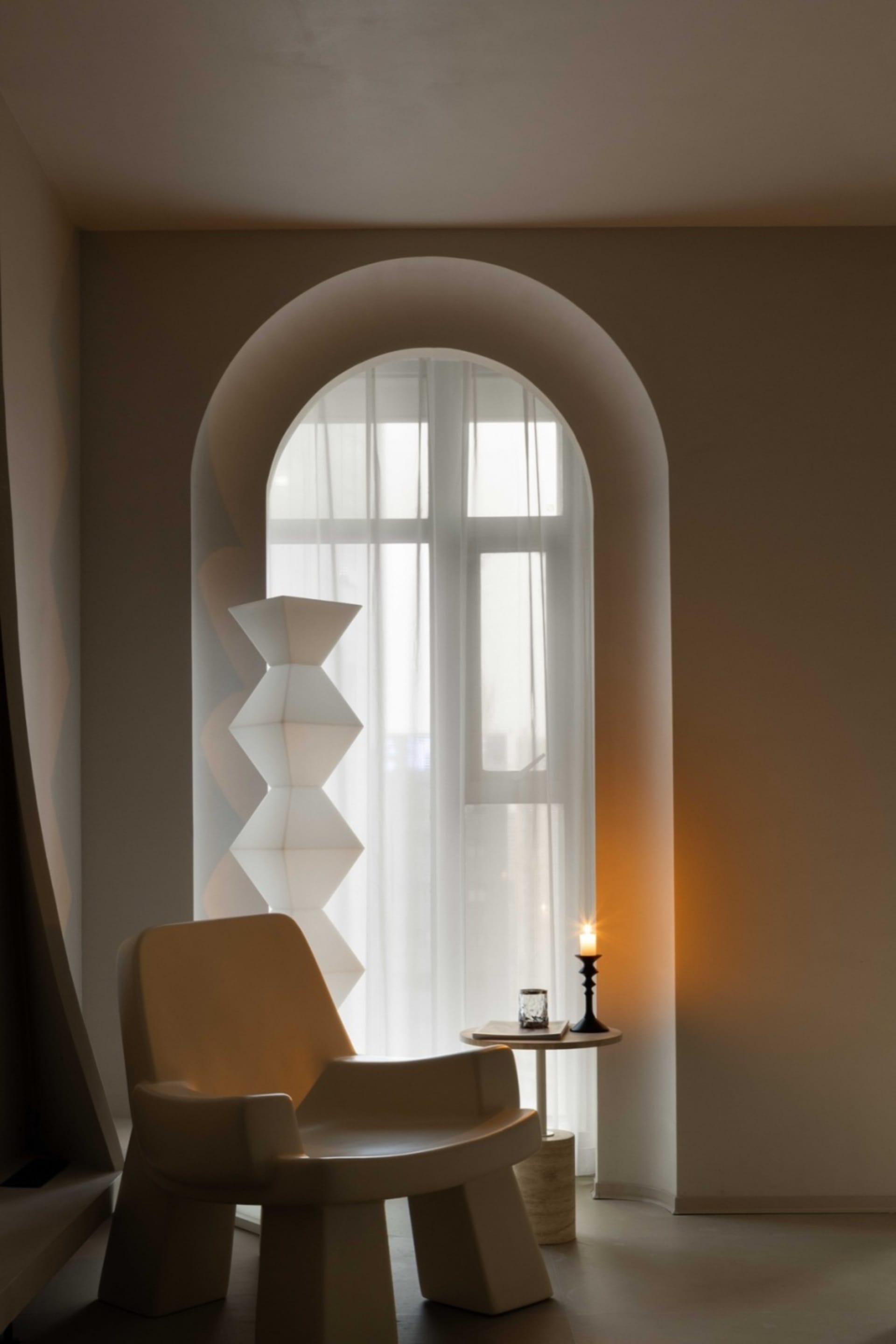 A minimalist living room with a large arched window featuring white curtains, a modern white armchair, and a small table with