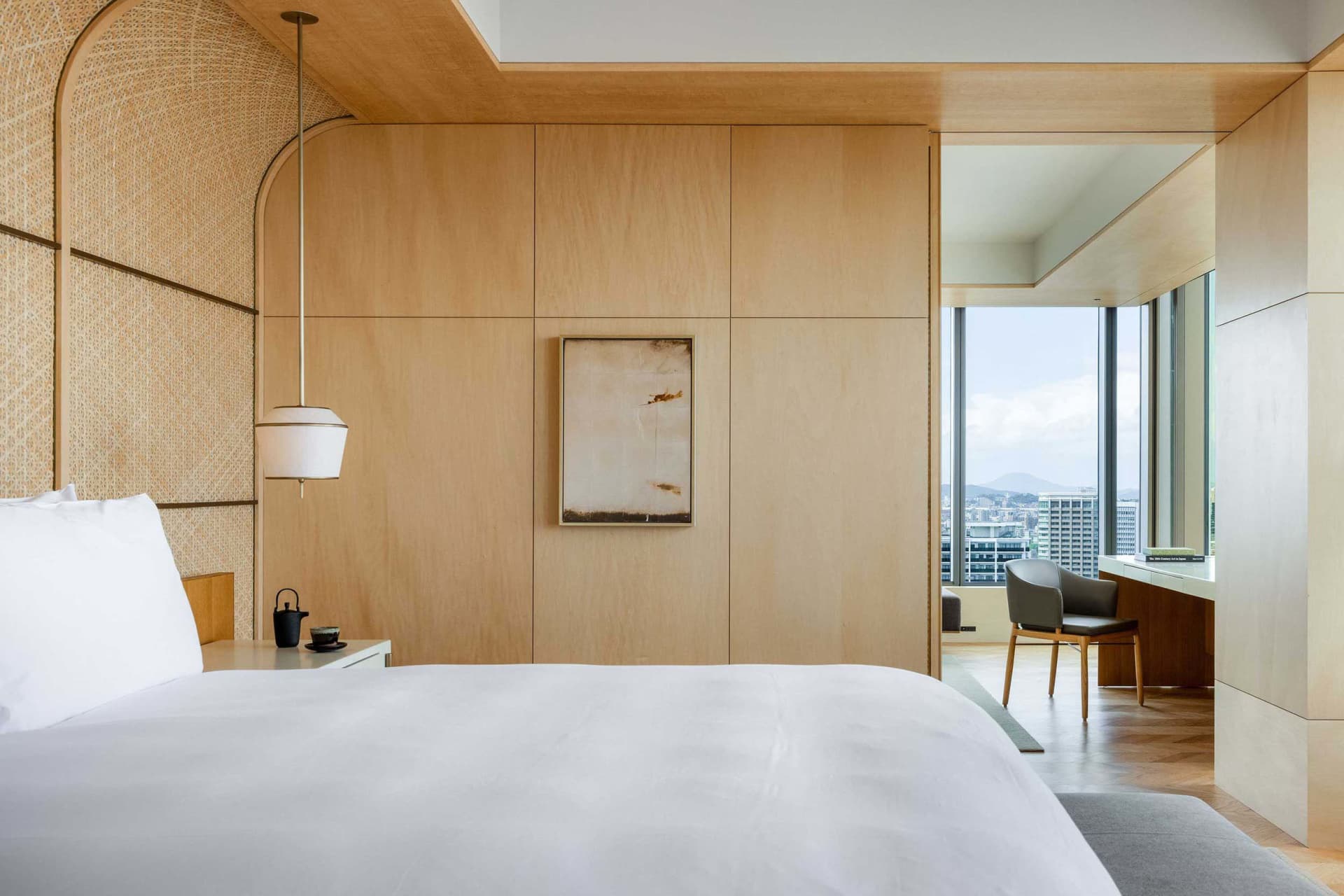 A modern bedroom with a bed, a chair, and a desk overlooking a cityscape.