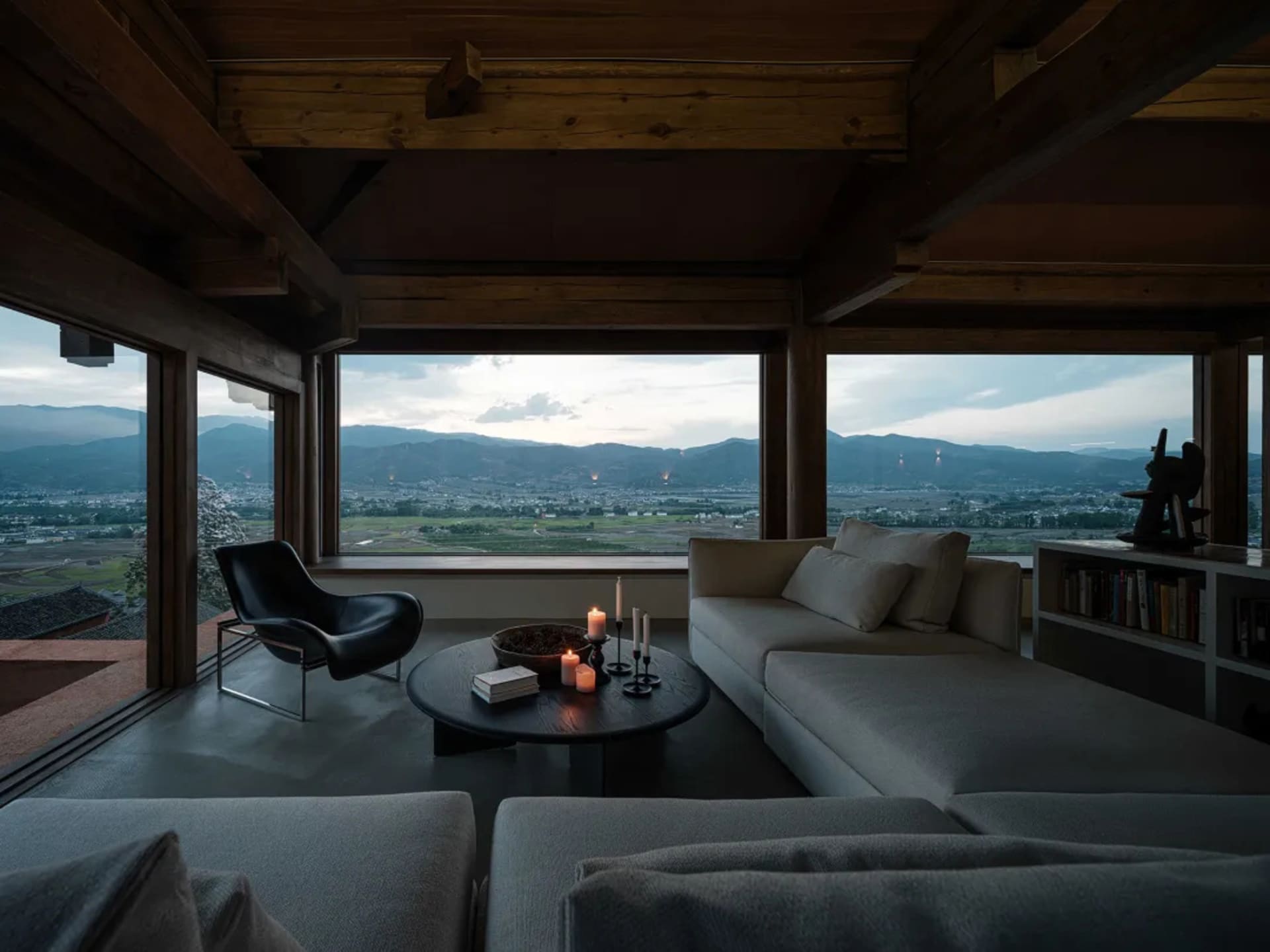A minimalist living room with a large window showcasing a scenic mountain view, featuring wooden beams, white sofas, and a co