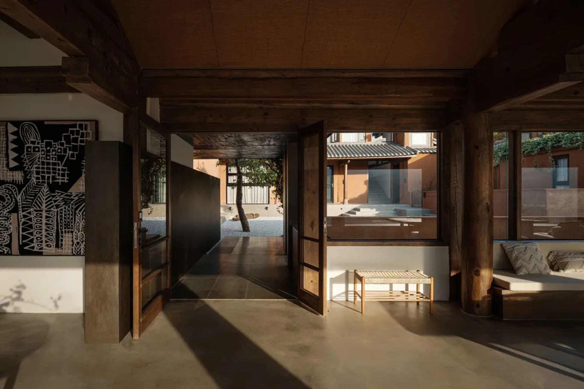 A modern living room with concrete flooring, wooden beams, and glass windows showcasing a serene outdoor view.