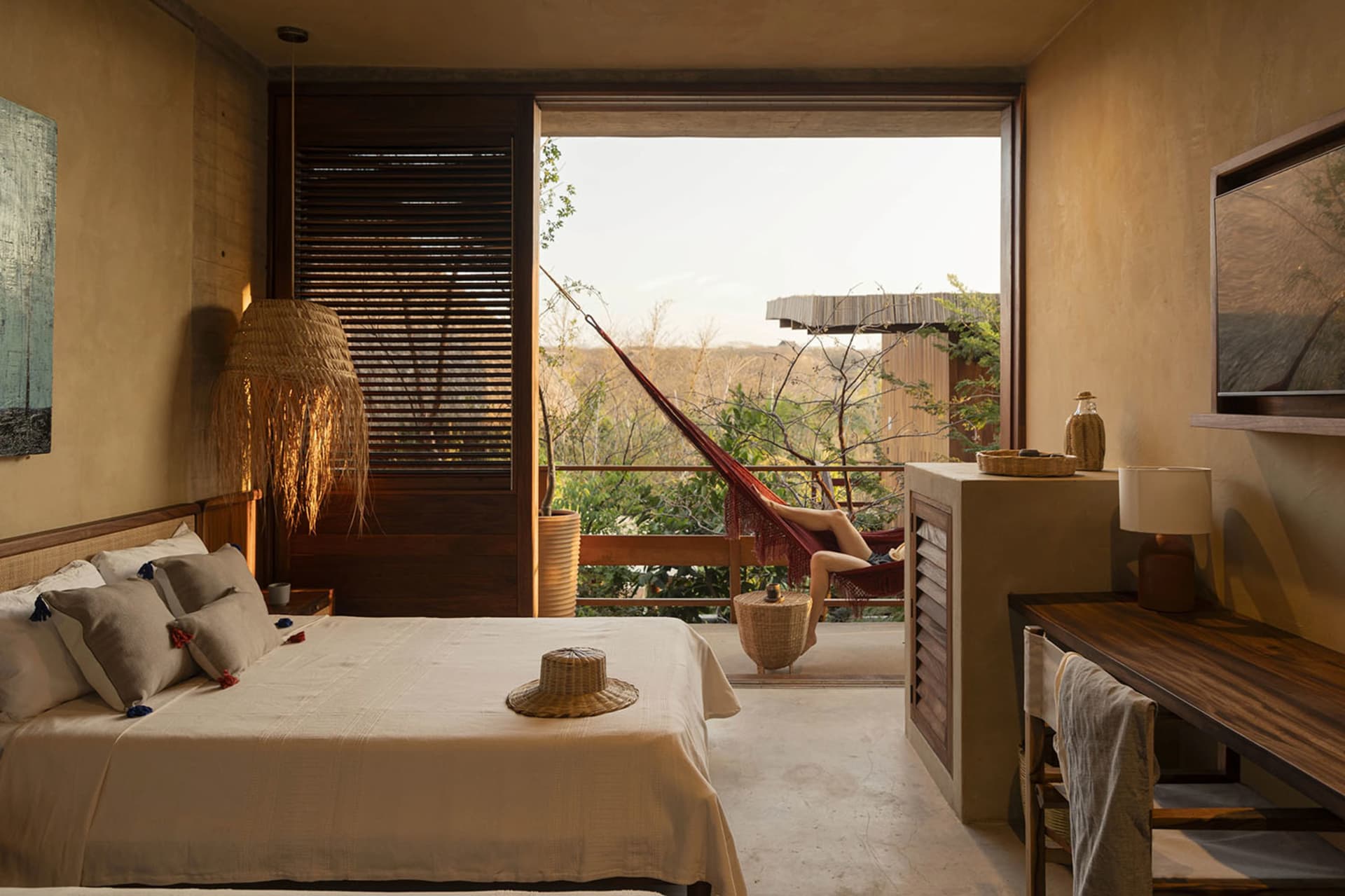 A minimalist bedroom with a bed, bedside table, and a hammock on the balcony overlooking trees.