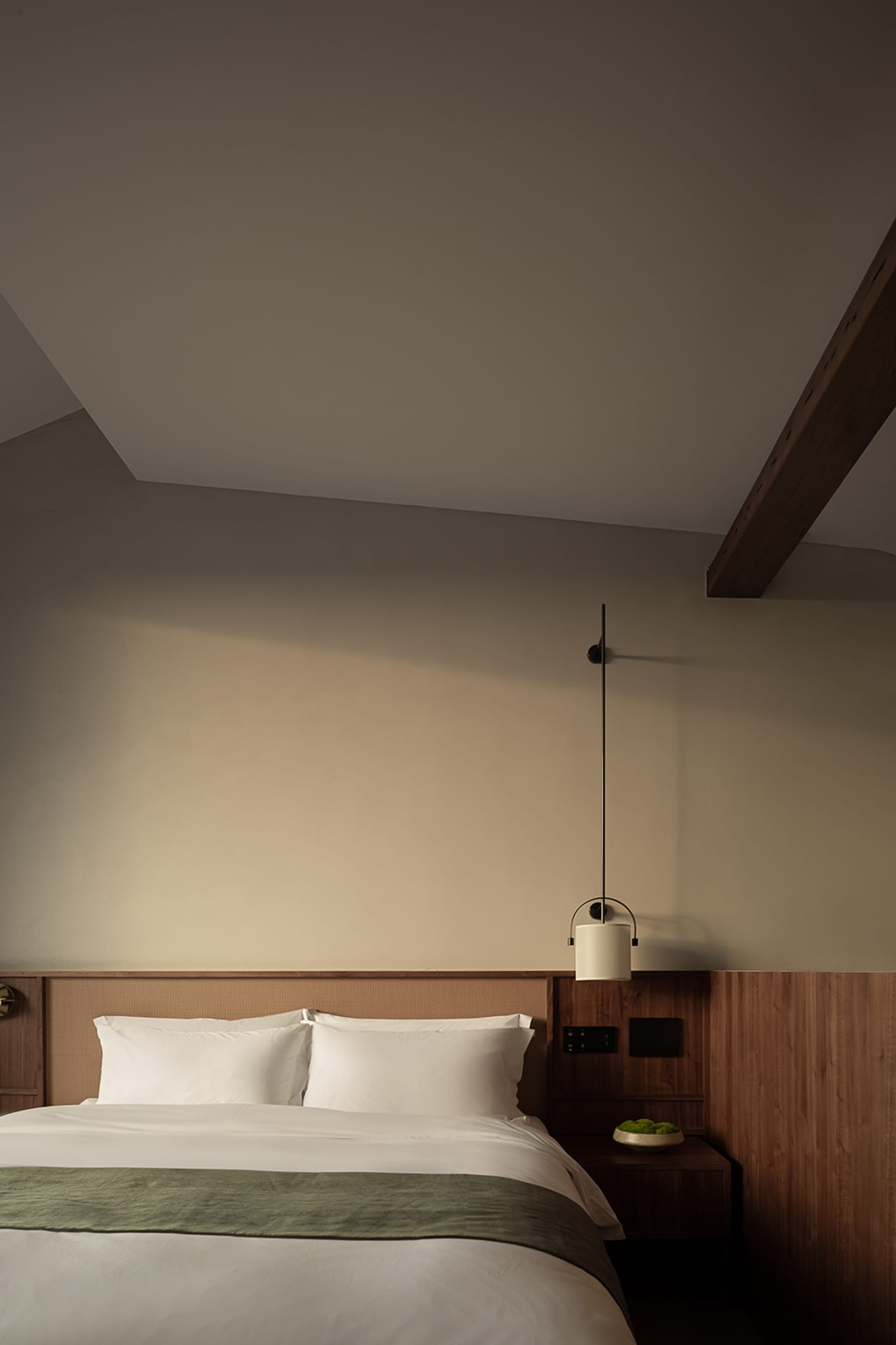 A minimalist bedroom with a wooden headboard and white bedding, featuring a hanging lamp.
