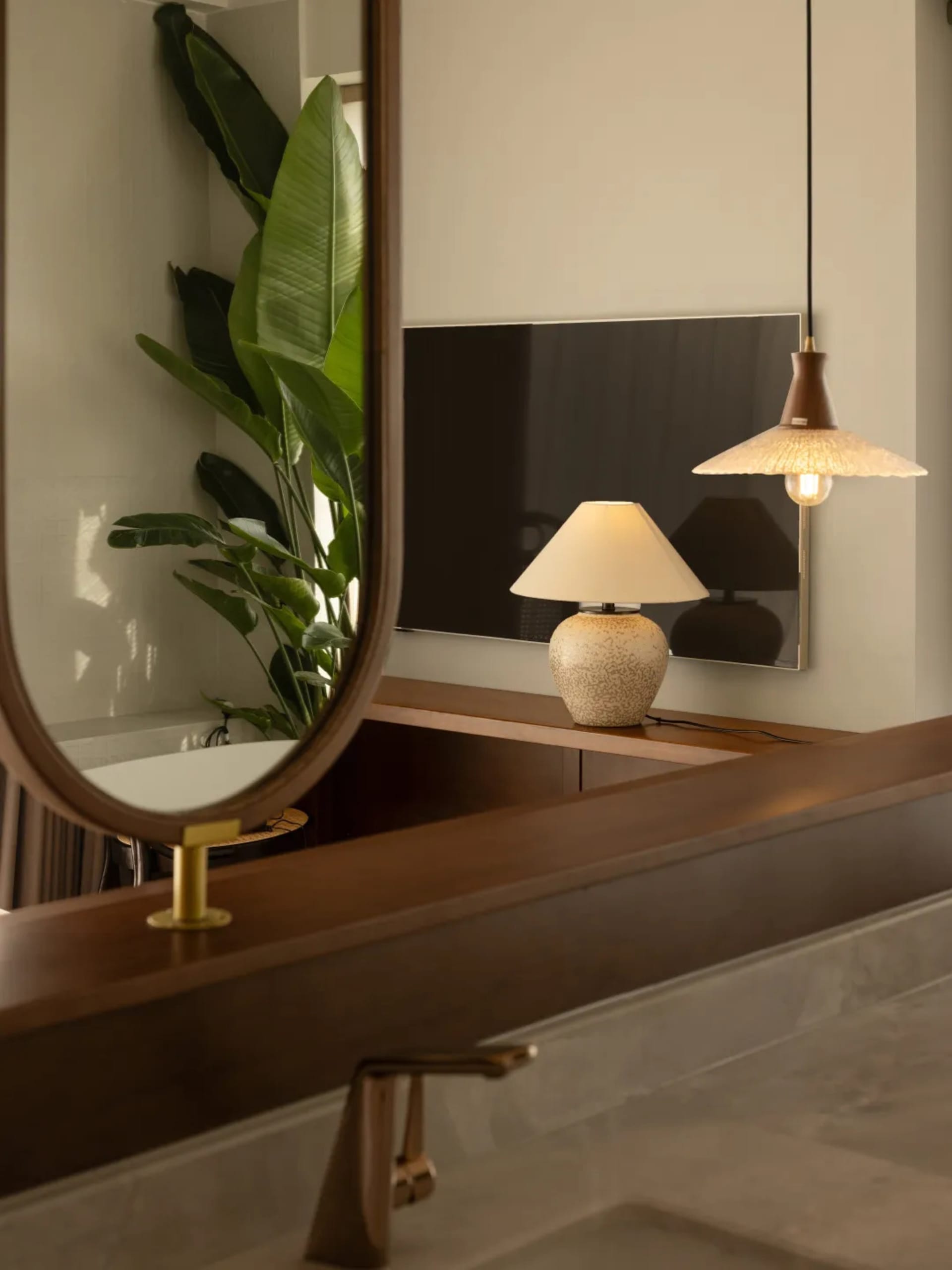 Modern bathroom interior featuring wooden vanity, ceramic lamp, glass pendant light, and green plant.