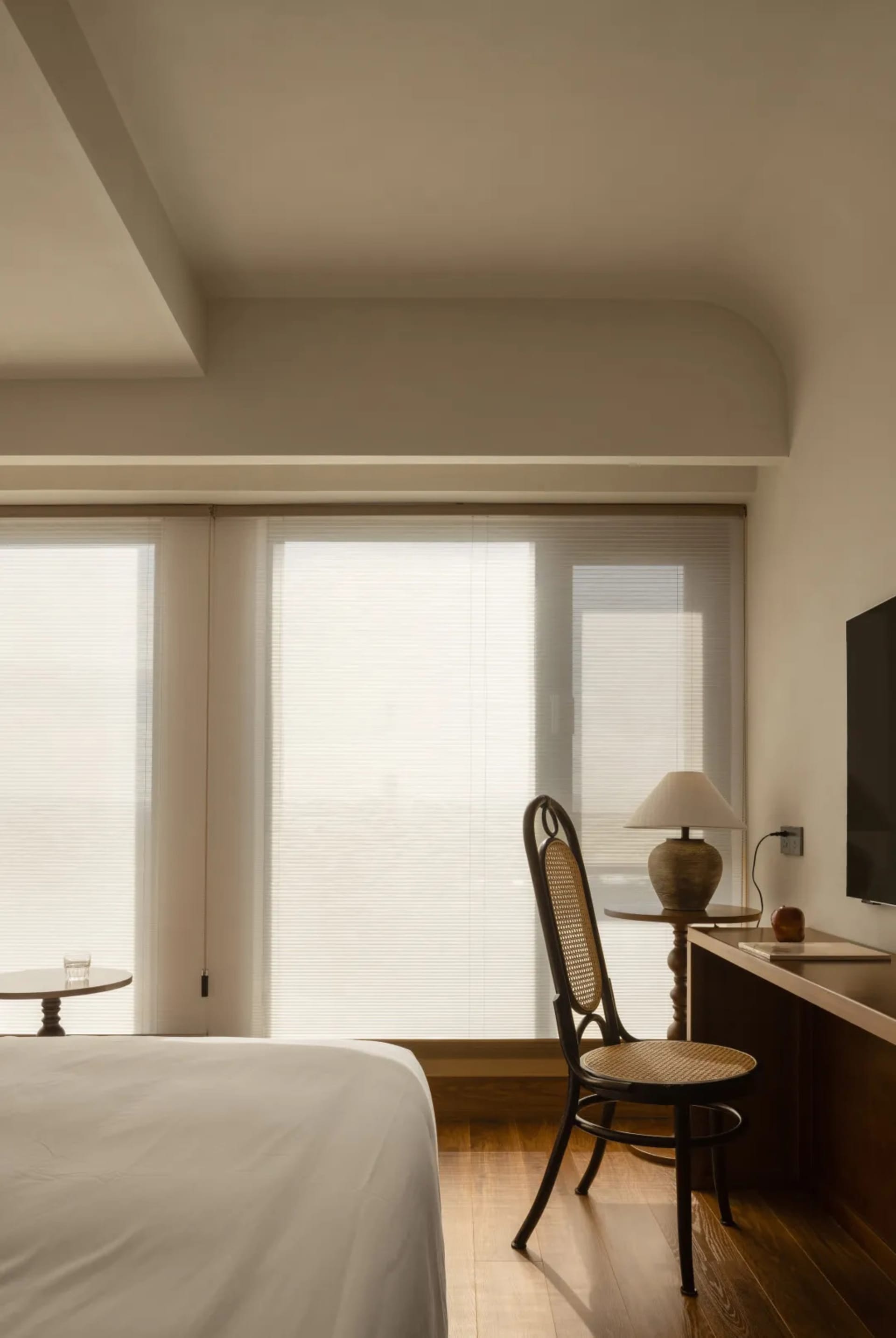 A modern minimalist bedroom featuring a bed with a white sheet, a wooden chair with a woven backrest, a lamp, and a televisio