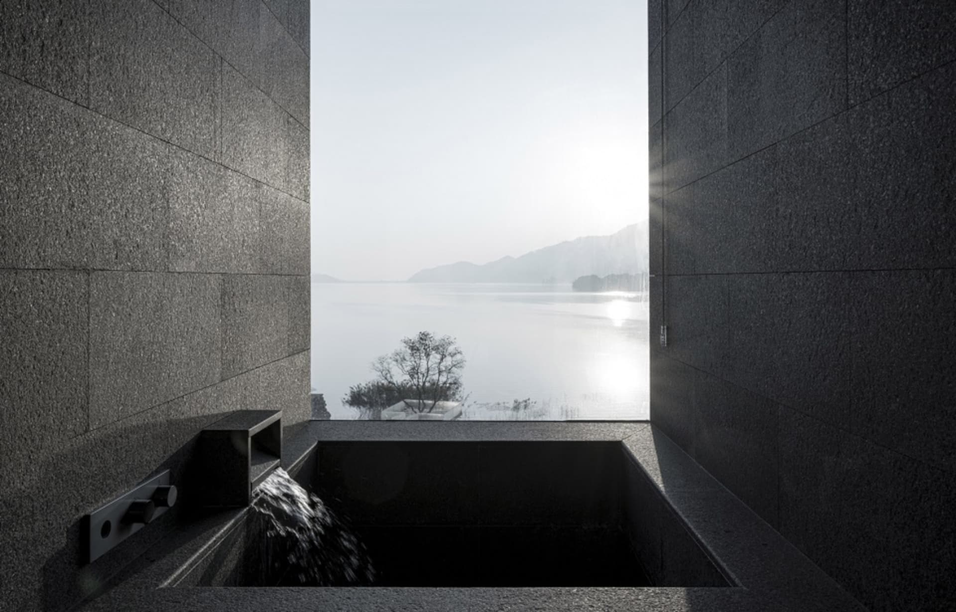 A modern stone relaxation bath overlooking a serene lake and mountains.