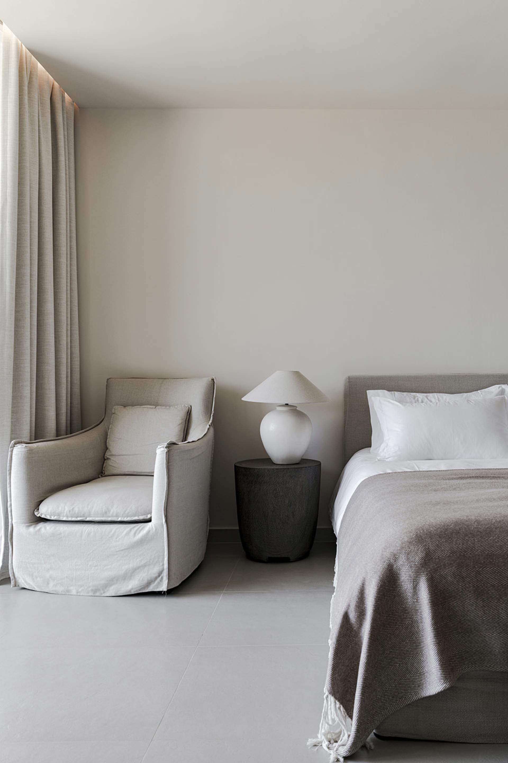 A minimalist bedroom with a bed, nightstand, lamp, and armchair in neutral tones.
