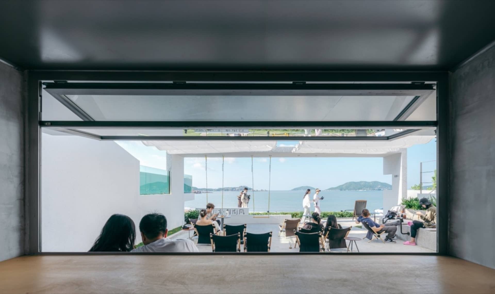 People sitting near a large glass window overlooking the ocean and mountains.