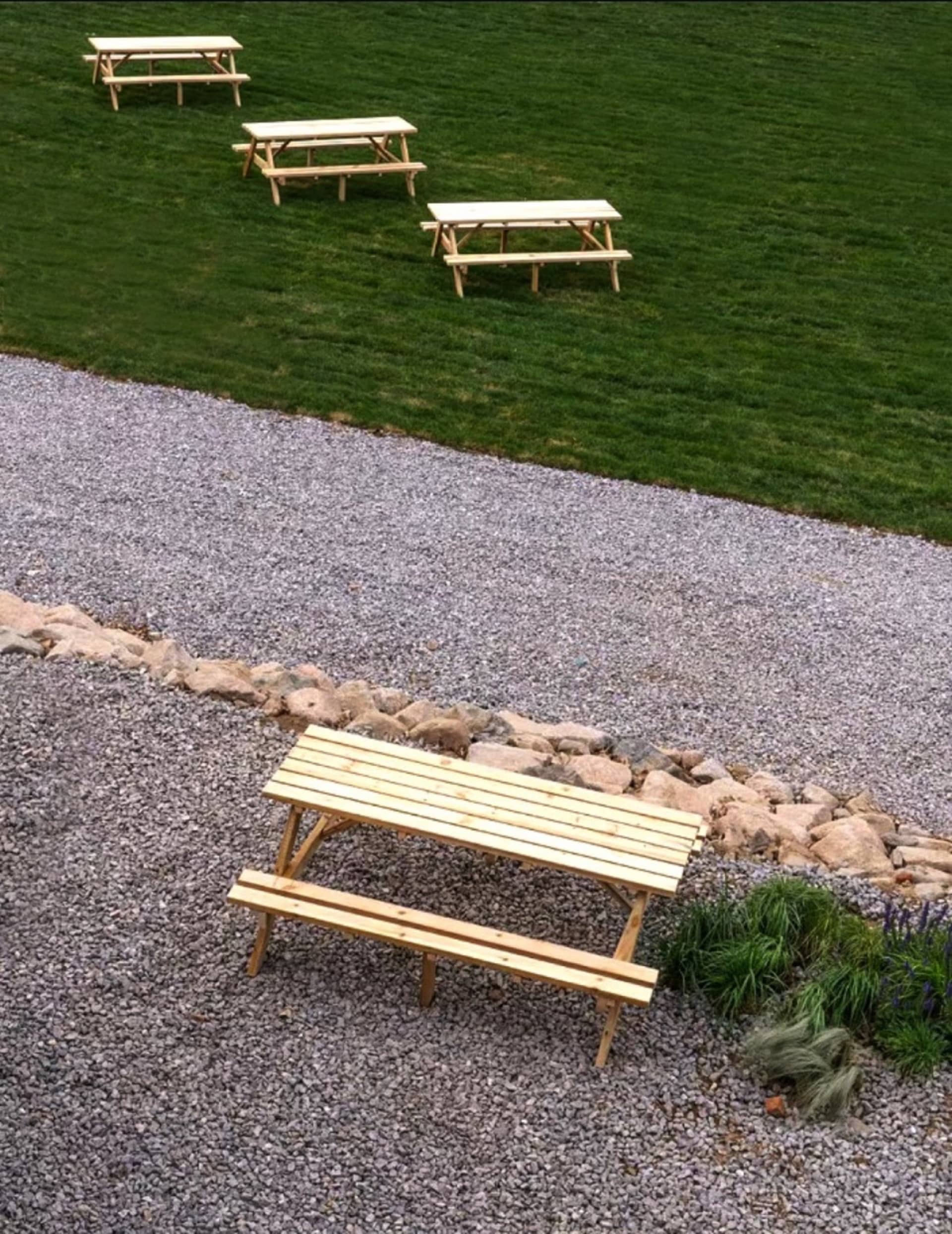 Four wooden picnic benches placed outdoors on a gravel surface surrounded by rocks and grass.