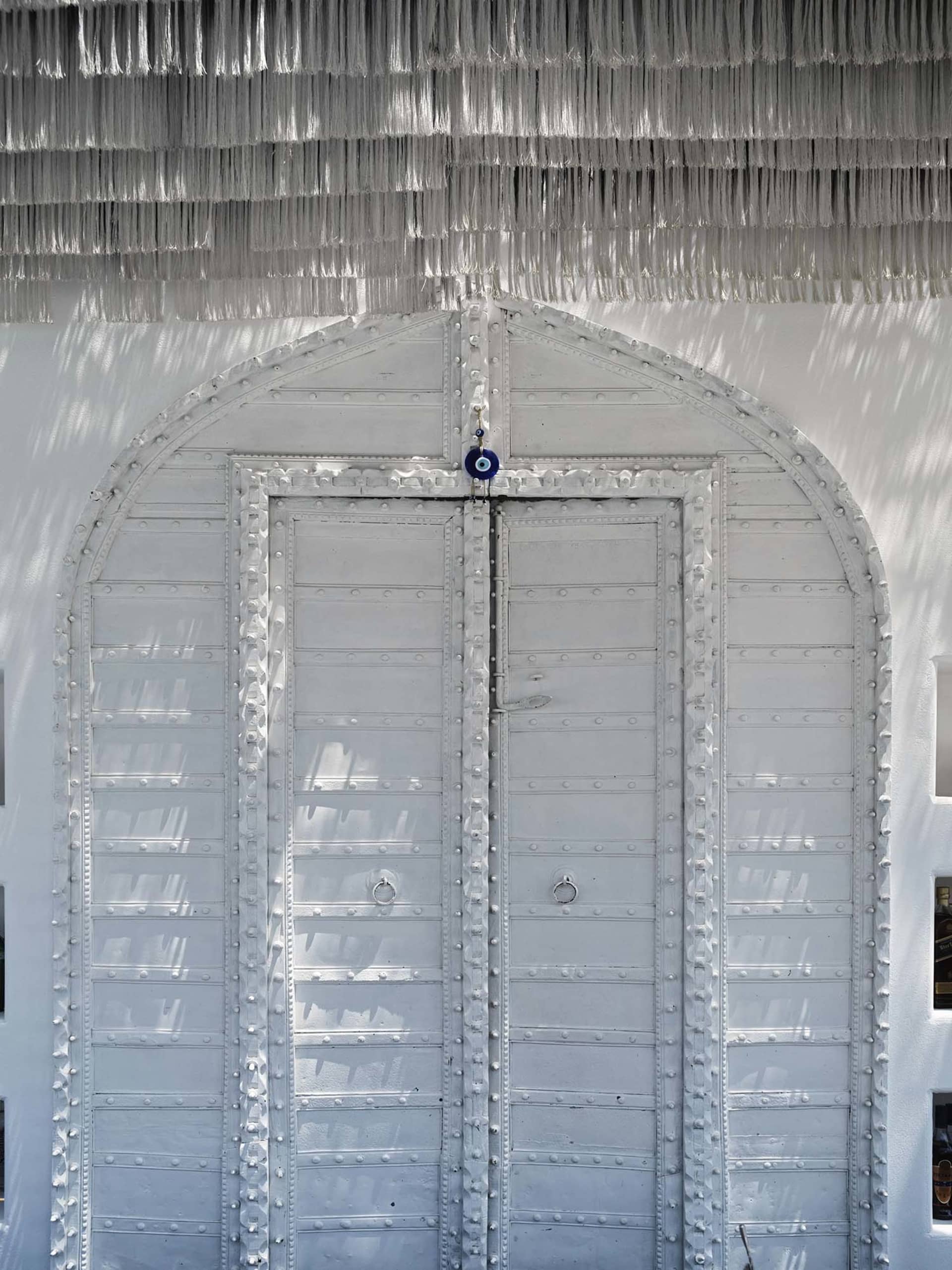 A white metal door with a decorative fringe on top, set against white walls with sunlight casting shadows.