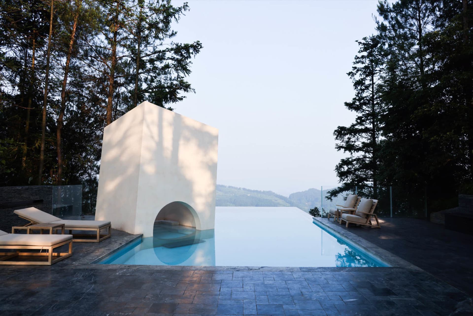 A minimalist concrete swimming pool with a cube-shaped structure and an infinity edge, surrounded by trees and overlooking di