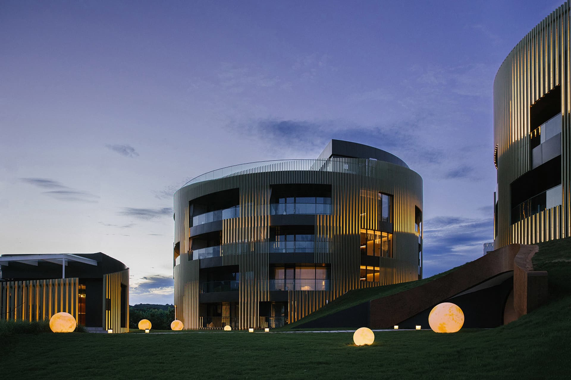A modern building with a metal-clad exterior illuminated at dusk with spherical light installations in the foreground.
