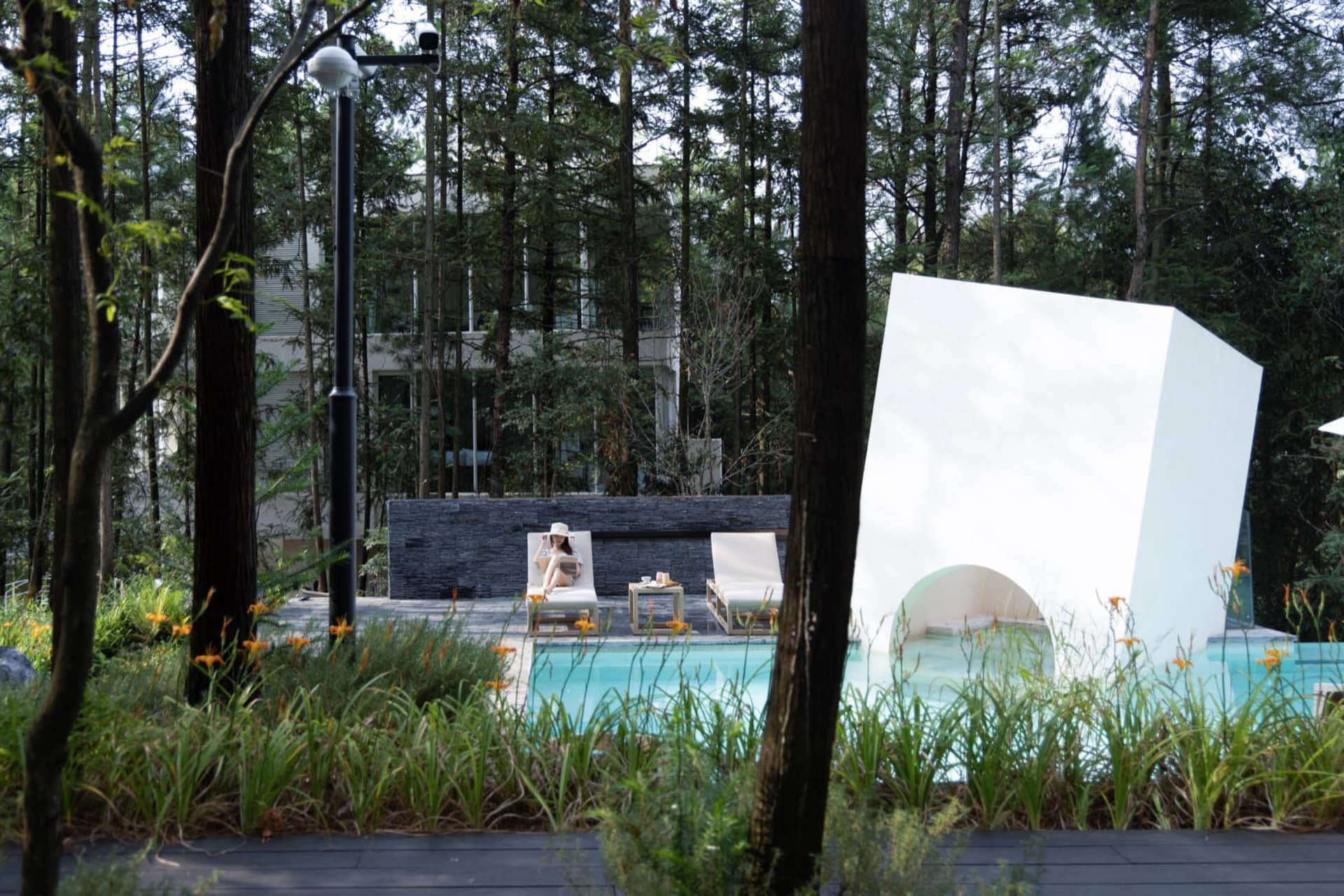 Modern residential exterior with a poolside relaxation space featuring wood, concrete, and glass materials.