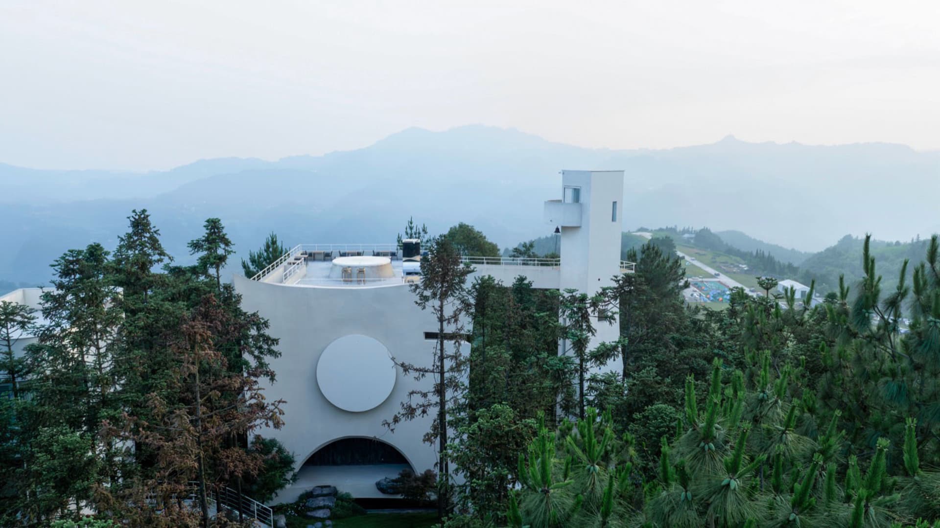 A modern white residential building with a white circular window and a glass entrance door surrounded by greenery.