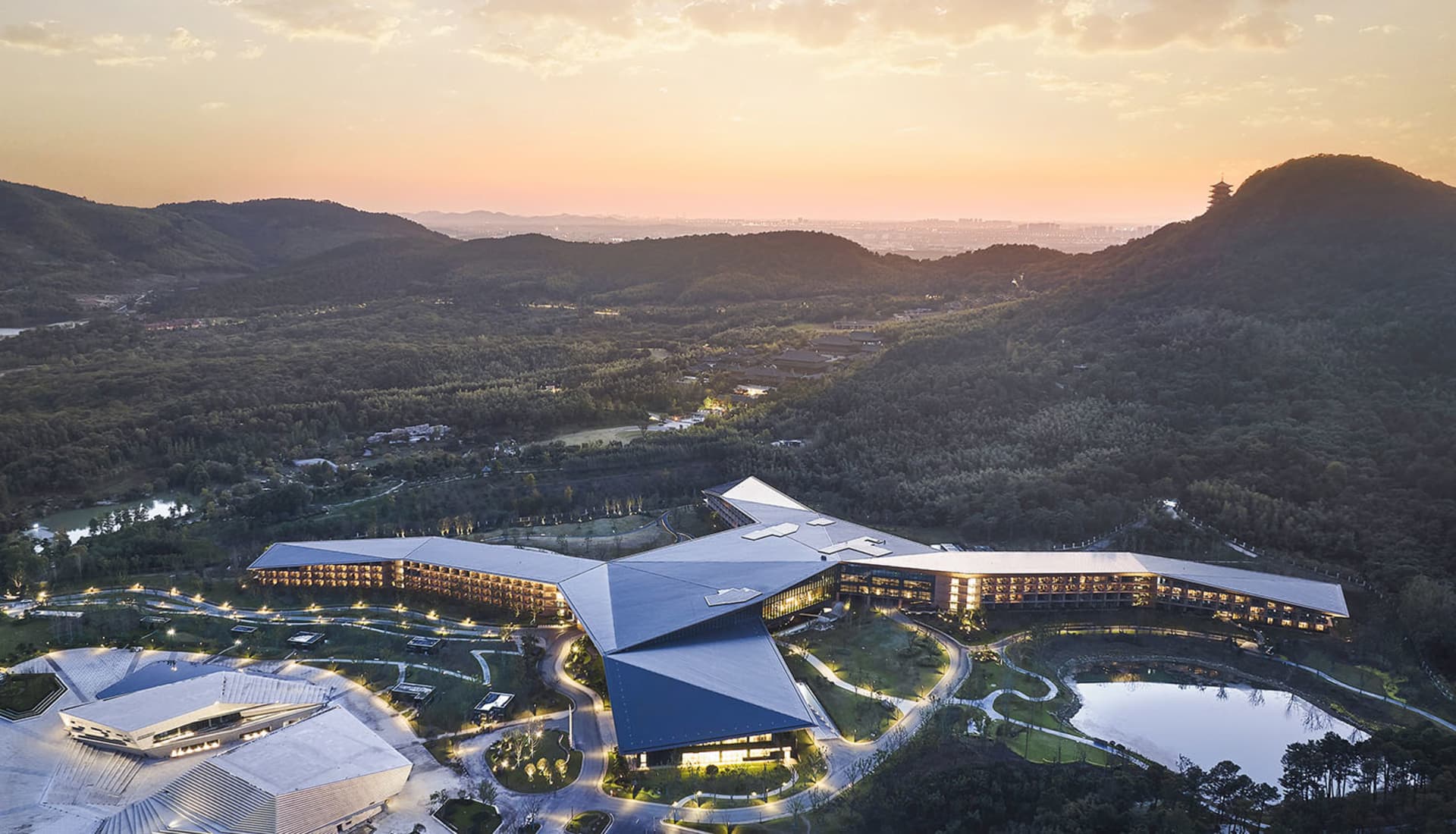 An aerial view of a modern hotel exterior featuring metallic, glass, and concrete materials, with a scenic landscape in the b