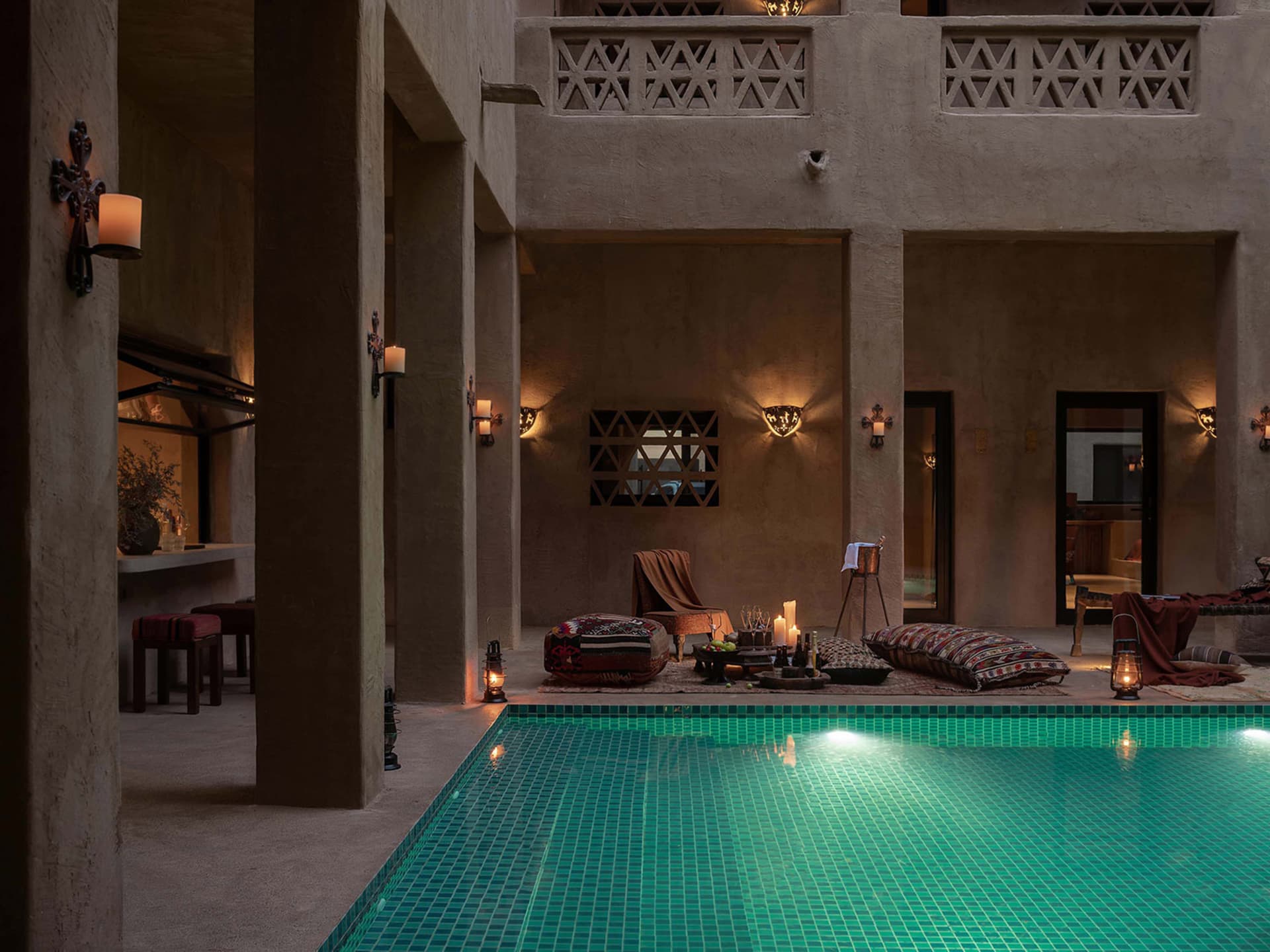 A modern residential lounge area with a poolside setup, featuring concrete walls and floors, soft furnishings, and warm light