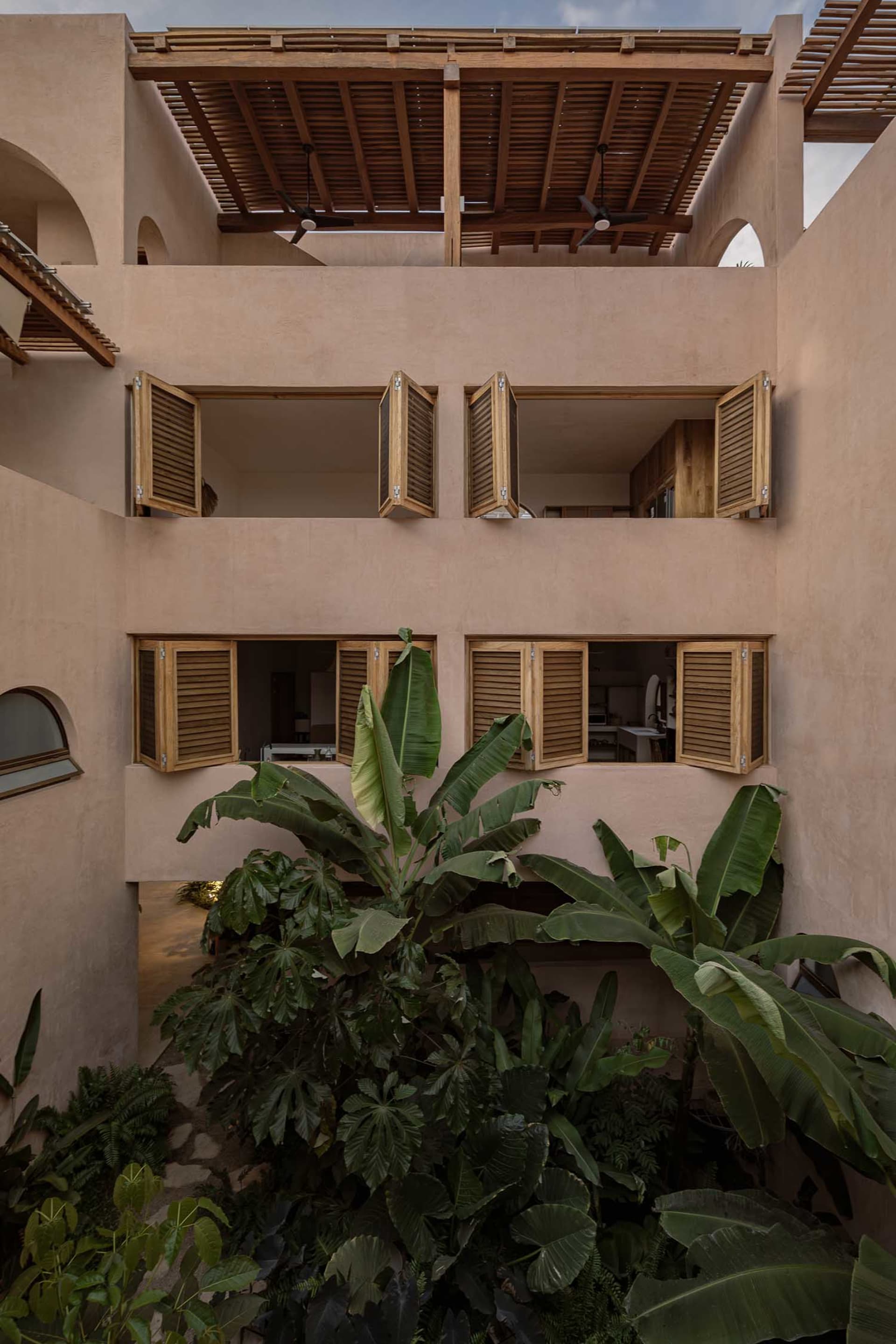 A clay-colored residential exterior with wooden shutters and a tropical garden.
