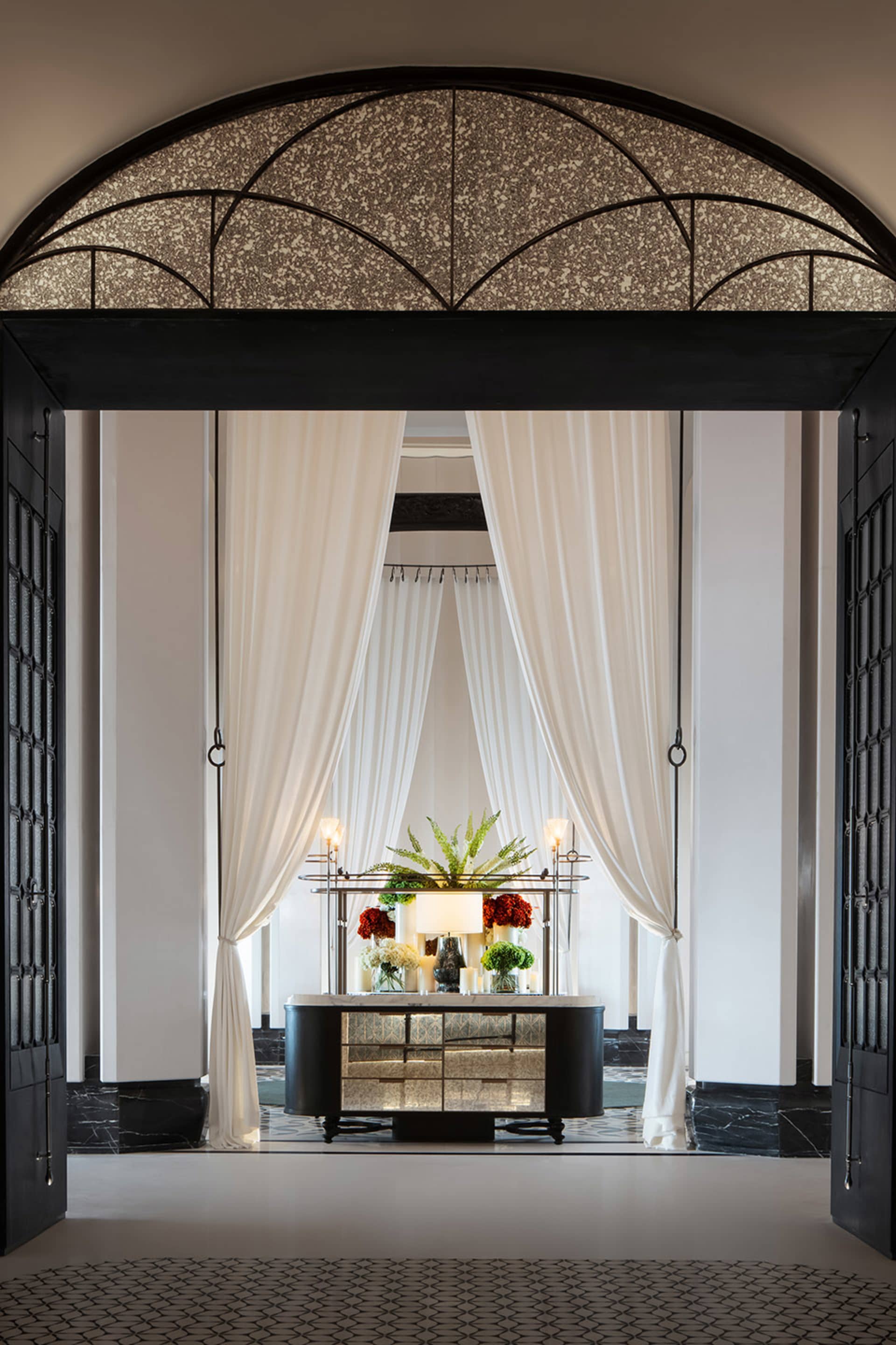 A luxury lobby featuring elegant white curtains with a decorative archway above the entrance, a table displaying flowers and 