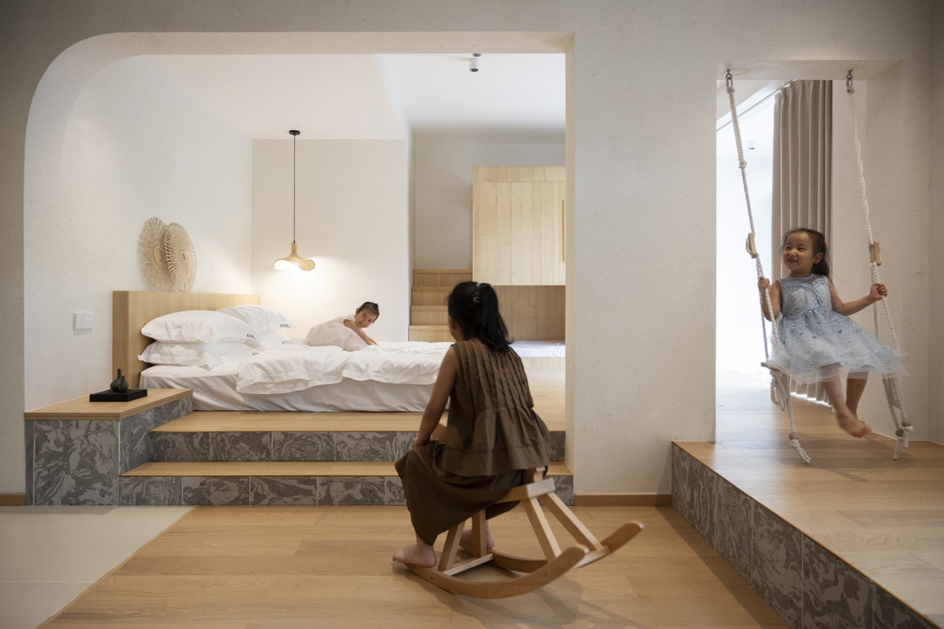 A modern playroom with warm tones, wooden and marble surfaces, and children playing on a wooden rocking horse and a swing.