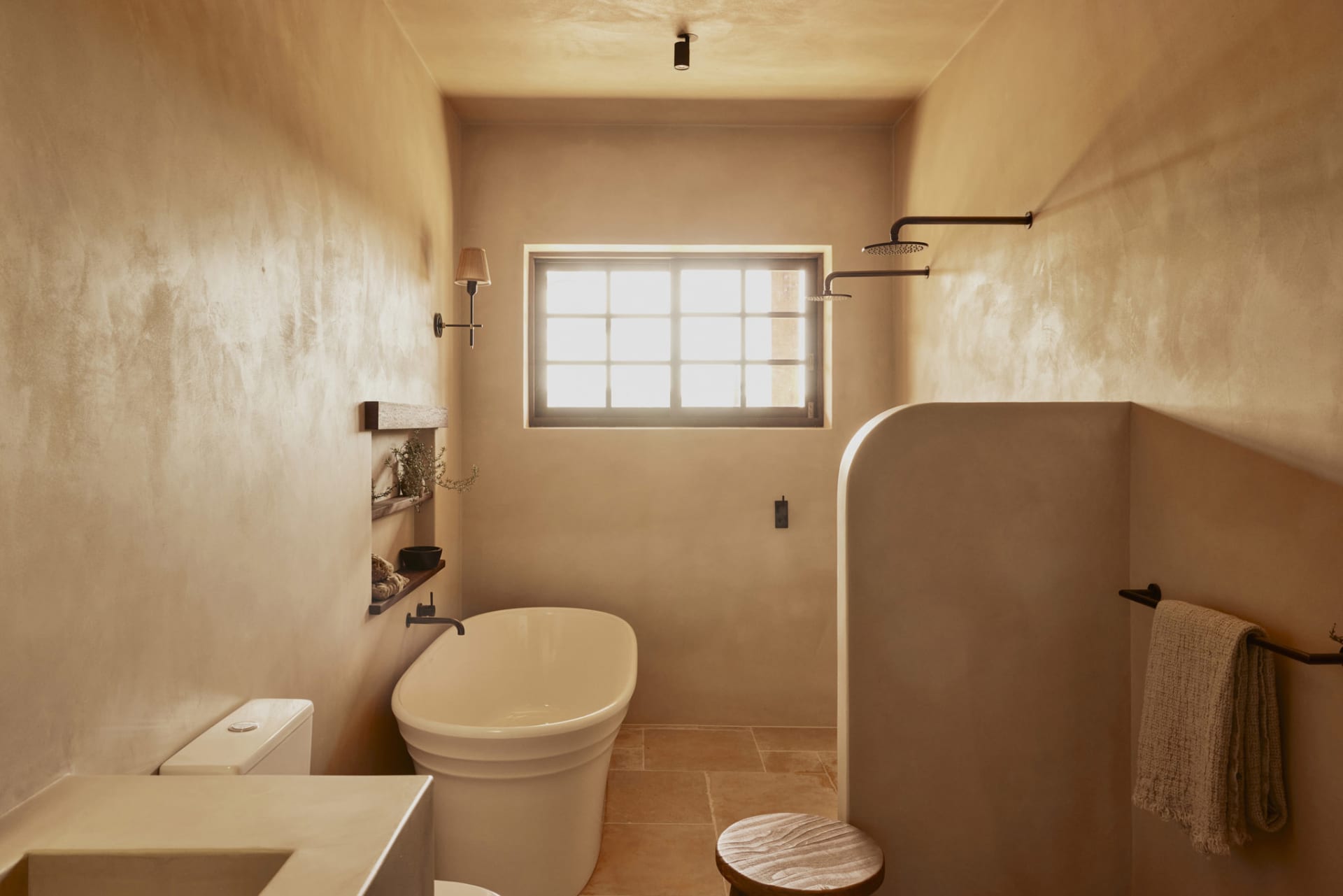 A minimalist bathroom with concrete walls, featuring a freestanding bathtub, a toilet, and a sink with a wooden stool. The ba