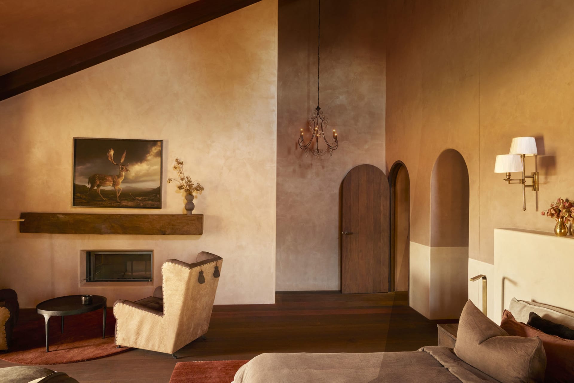 A modern bedroom with a warm color palette featuring a plaster accent wall, a chandelier, and a wooden bed.
