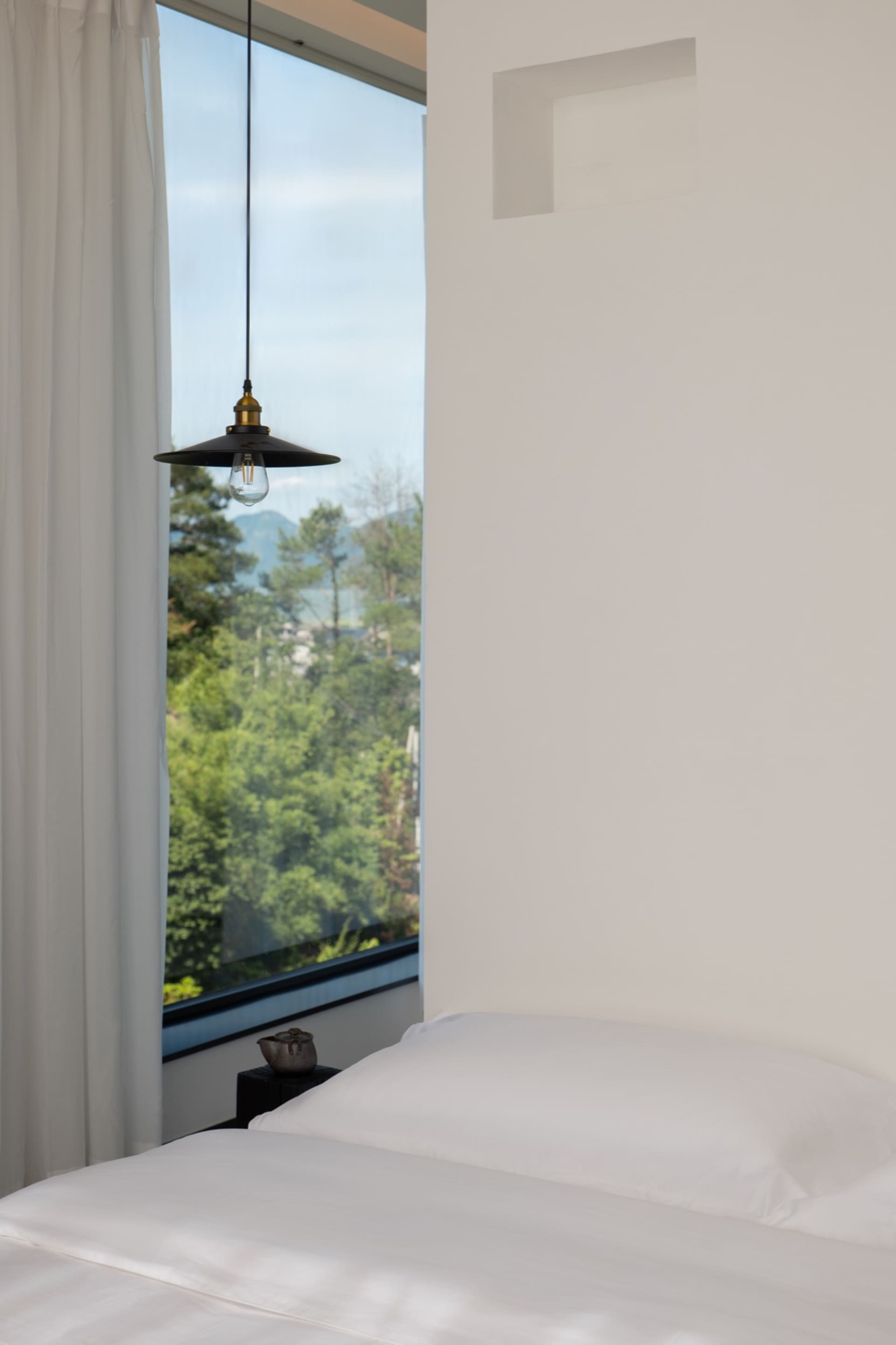 A modern white bedroom with a large window overlooking green trees and mountains, featuring a minimalist design with white be