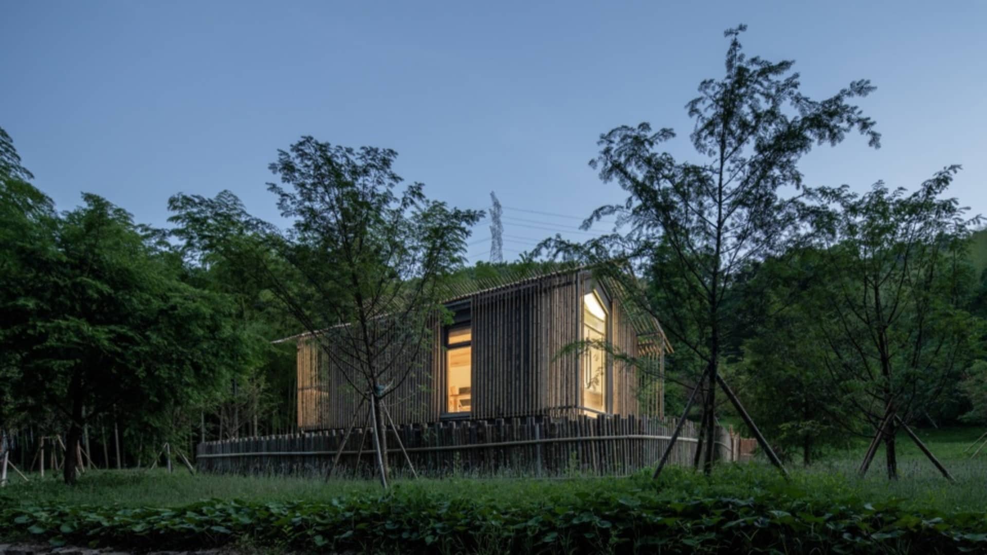 A minimalist building constructed primarily of wood, featuring a single illuminated window.