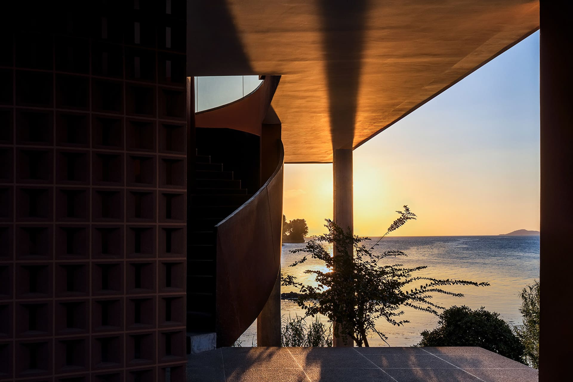 A modern concrete facade overlooking the ocean during sunset, with a plant in the foreground.